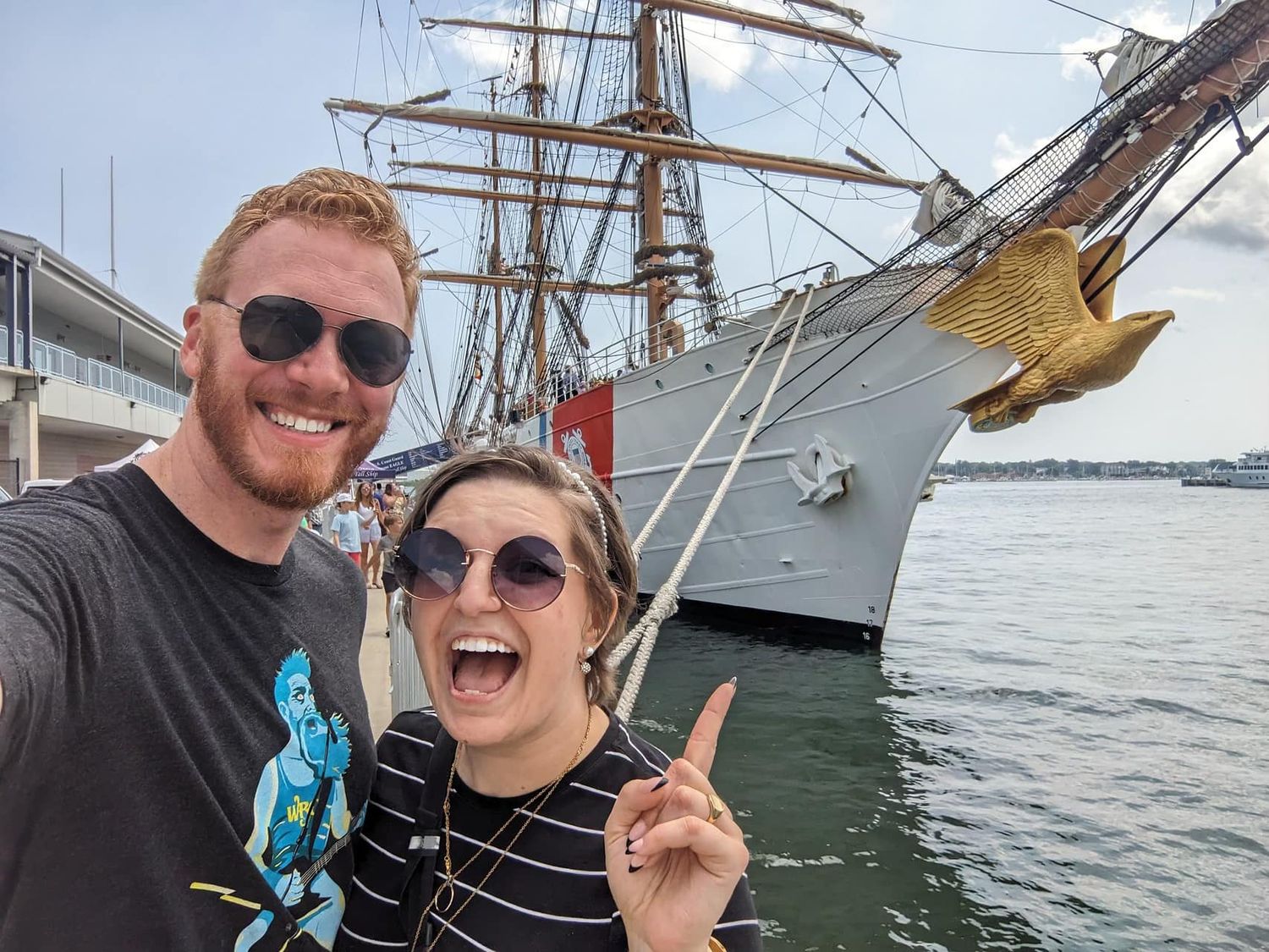 Smiling couple taking a selfie in front of a tall ship docked at a harbor pier.