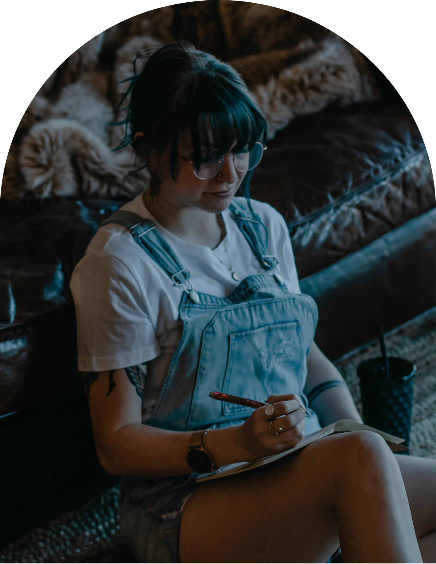 Young woman in denim overalls writing in a notebook while sitting on a couch indoors.