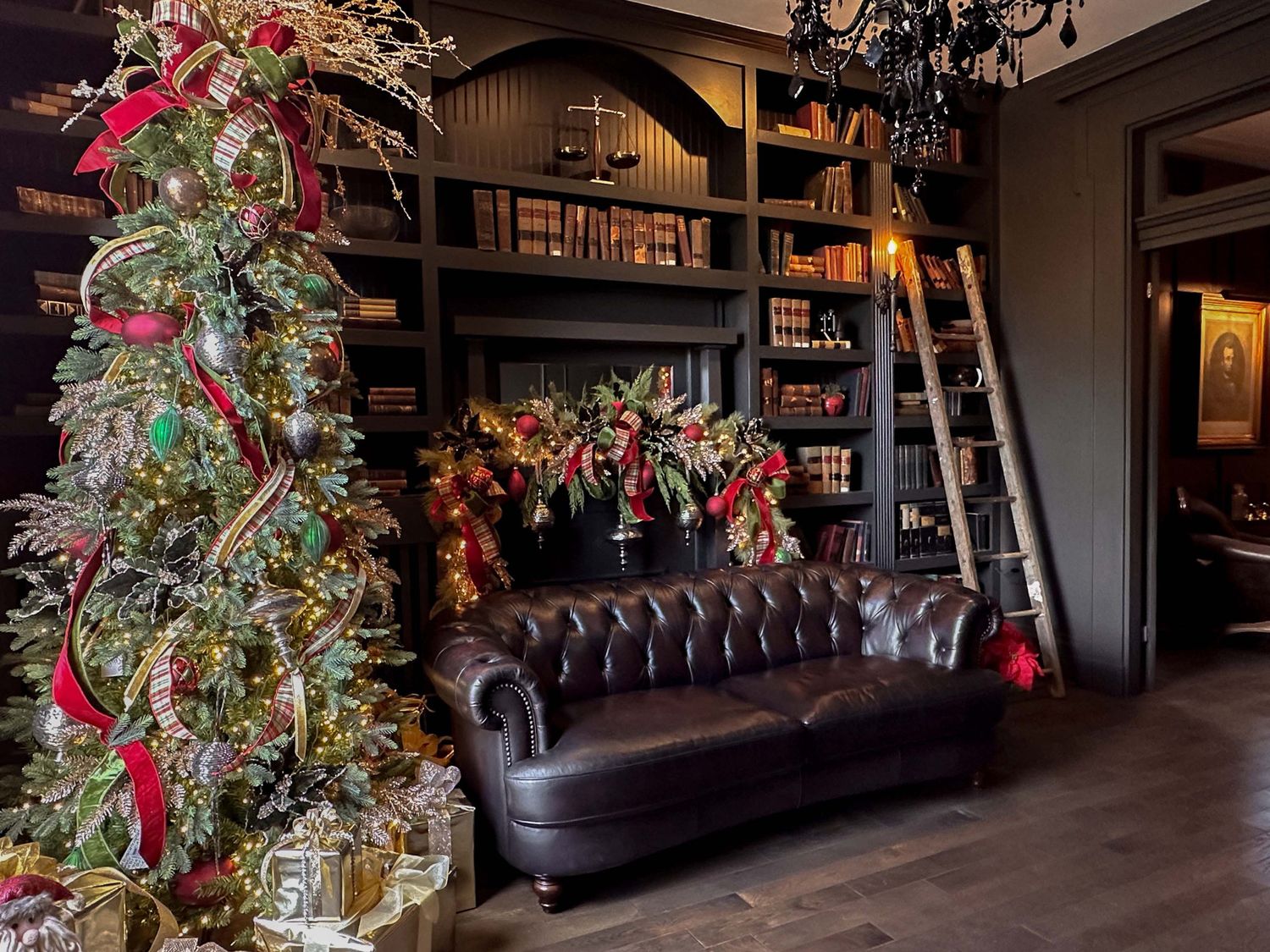 Elegant library with dark bookshelves, leather Chesterfield sofa, rolling ladder, and festive Christmas tree with red ribbons.