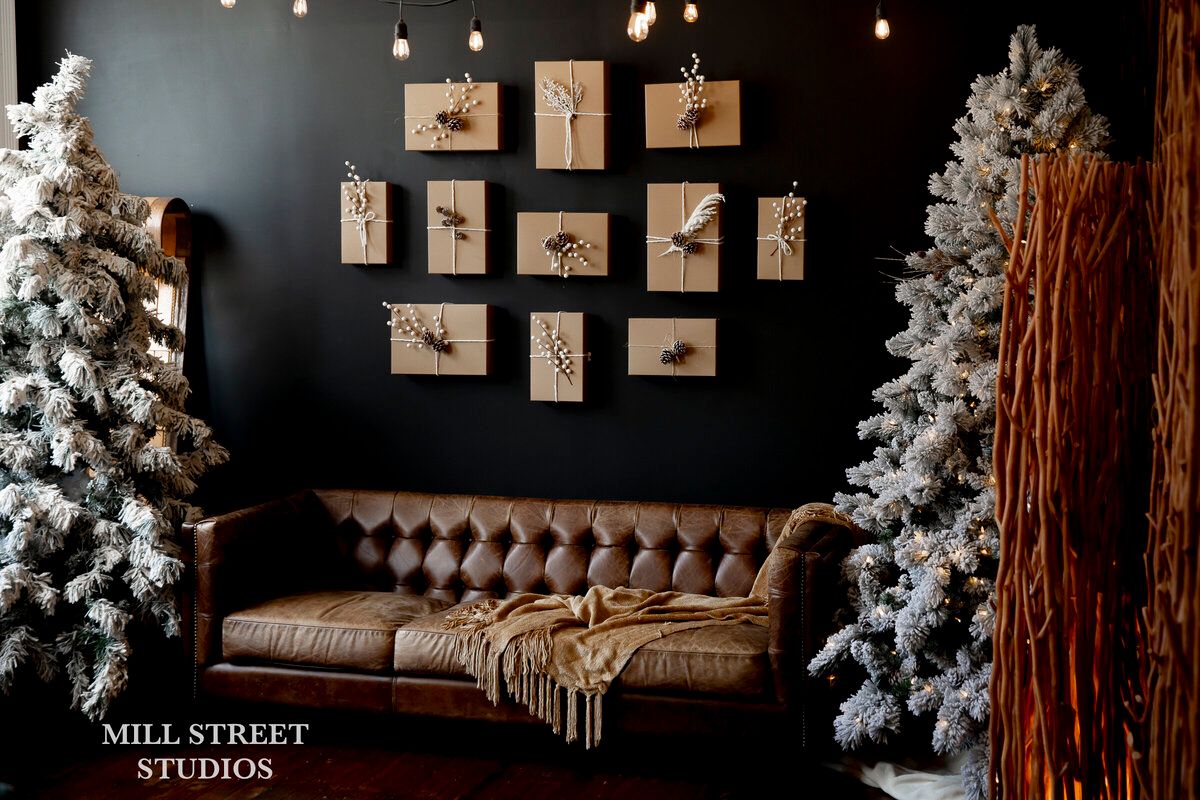 Cozy holiday studio with flocked Christmas trees, leather Chesterfield sofa, and kraft paper wall art on dark walls.