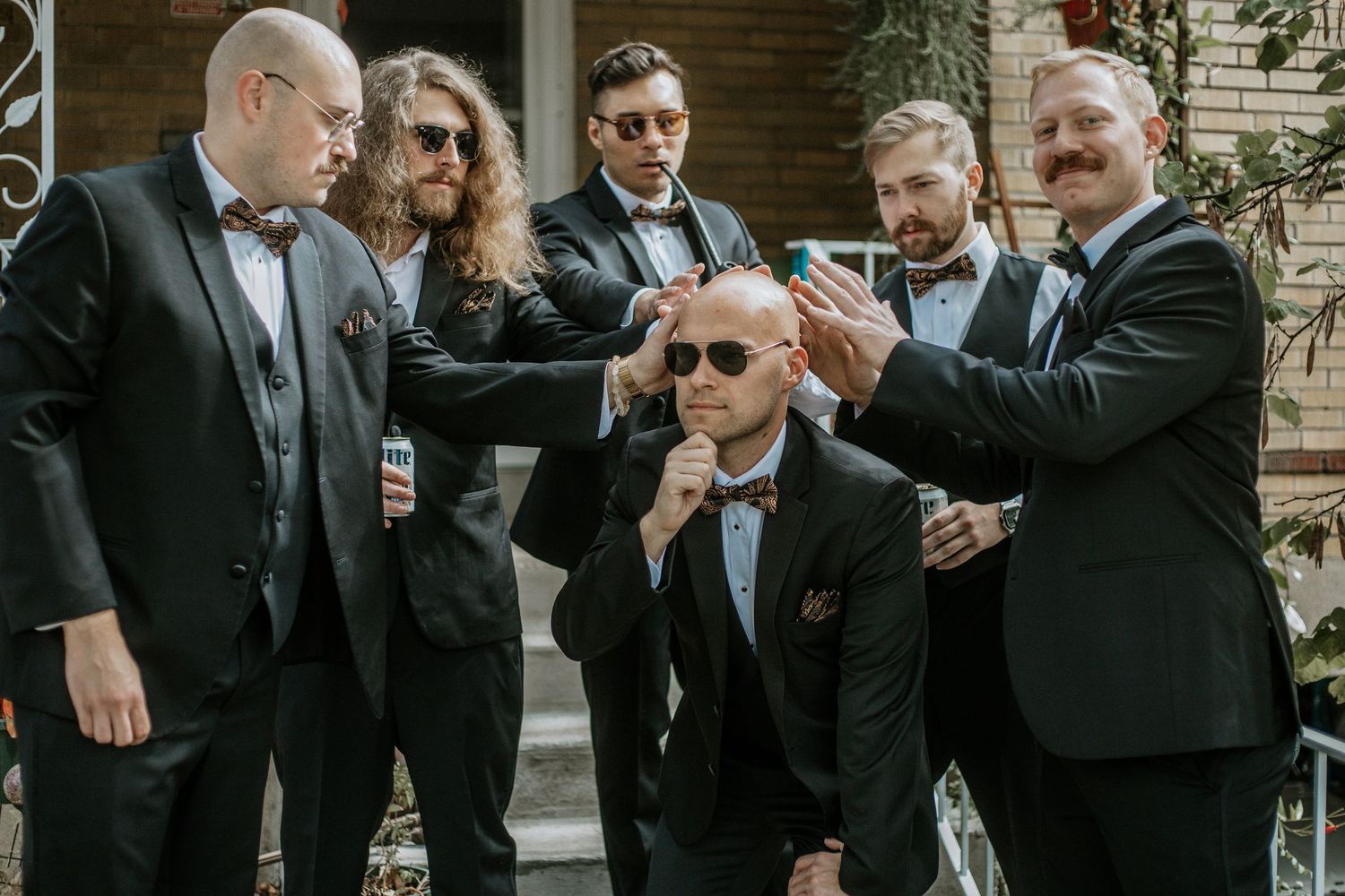 Six groomsmen in black tuxedos with bow ties pose together outdoors, adjusting sunglasses in a fun wedding party photo.