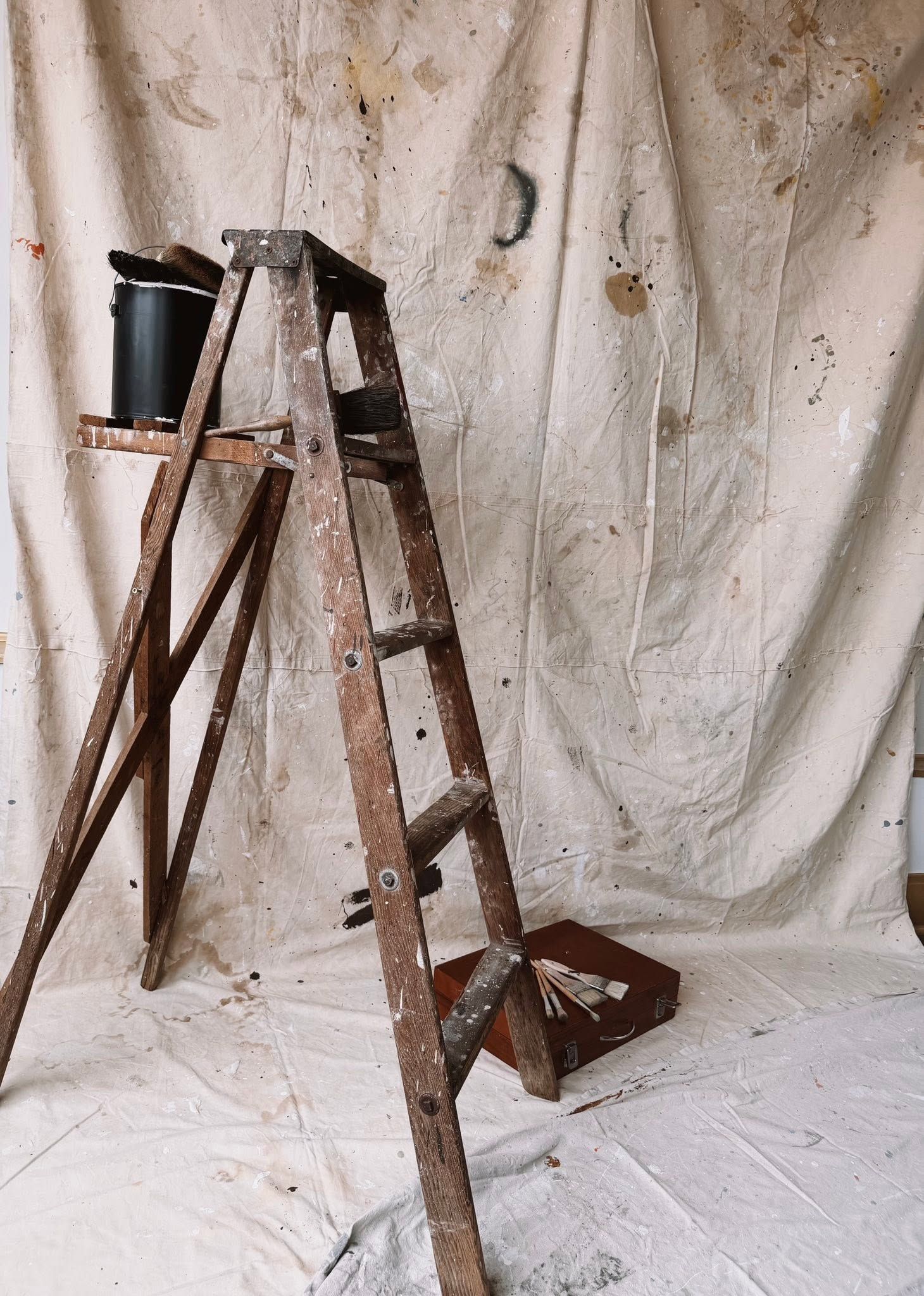 Weathered wooden A-frame ladder with paint bucket against stained canvas drop cloth in artist studio.