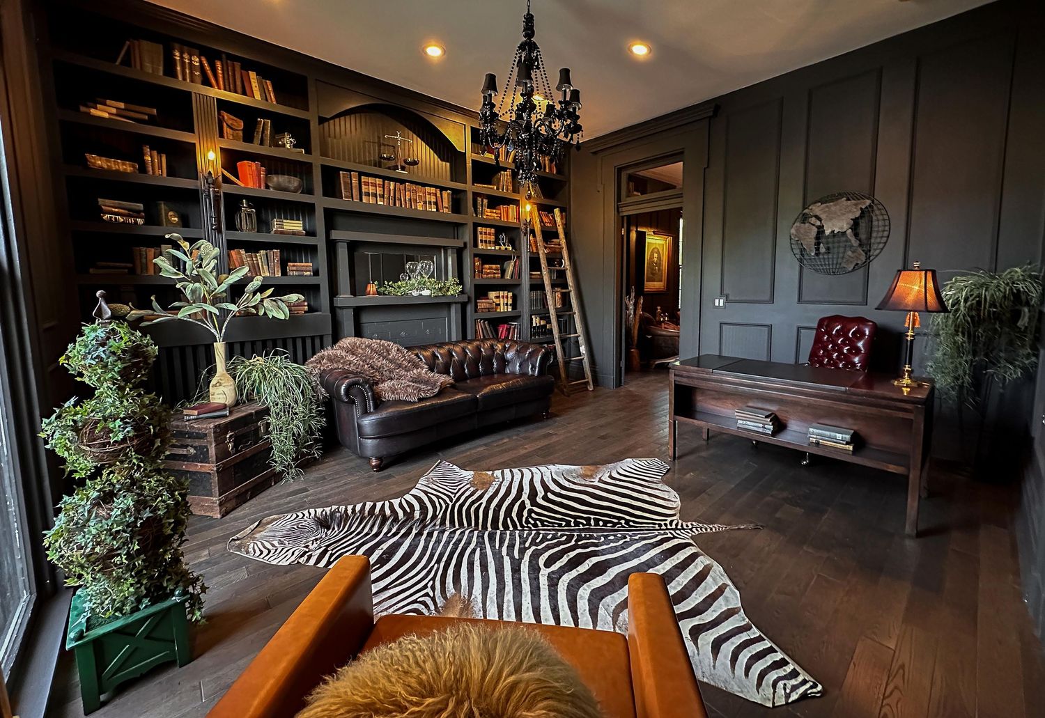 Dark moody library with zebra rug, leather Chesterfield sofa, built-in bookshelves, rolling ladder, and chandelier.
