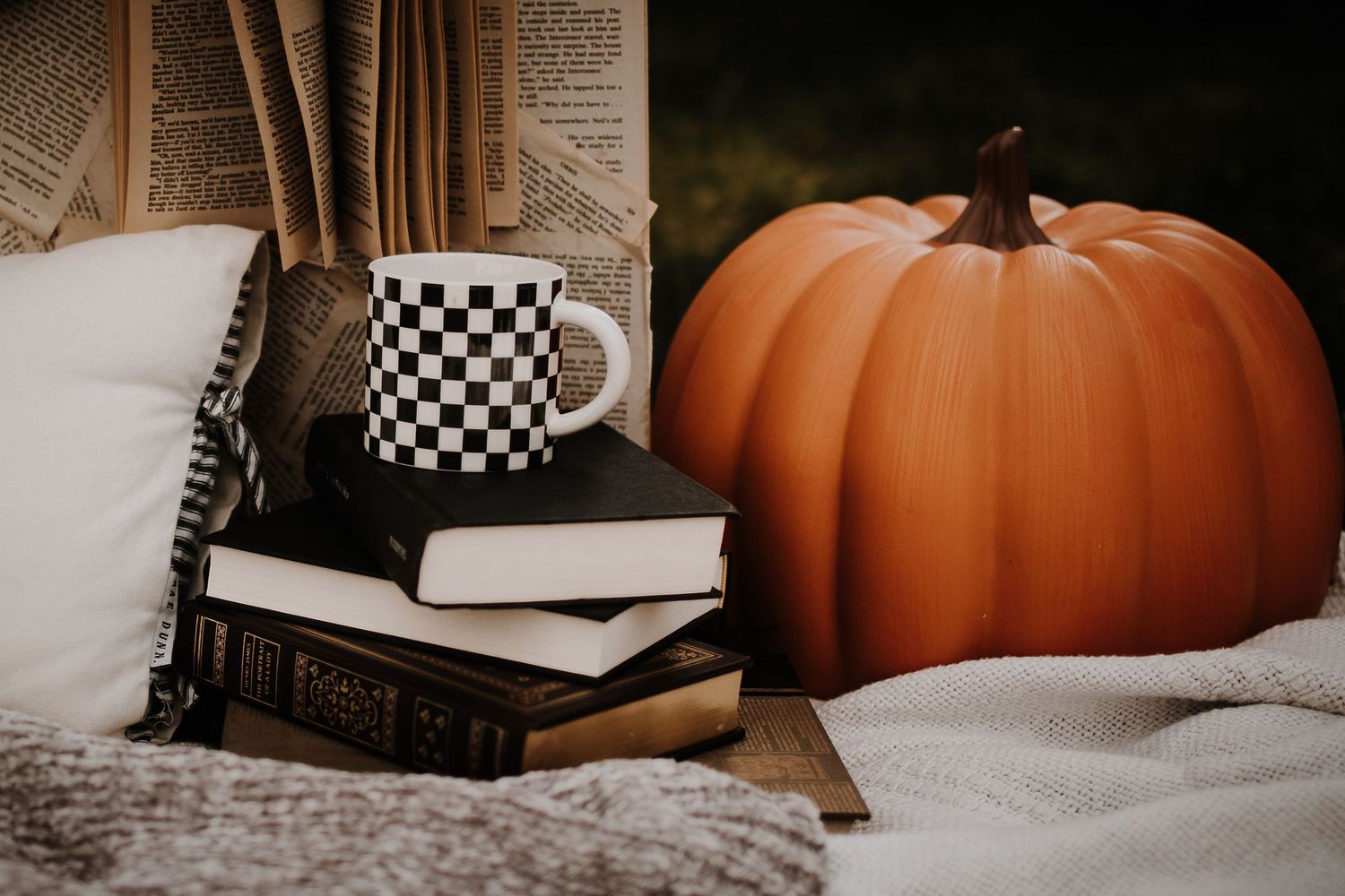 A cozy autumn vignette featuring a checkered mug on stacked books beside a large orange pumpkin with soft textiles.