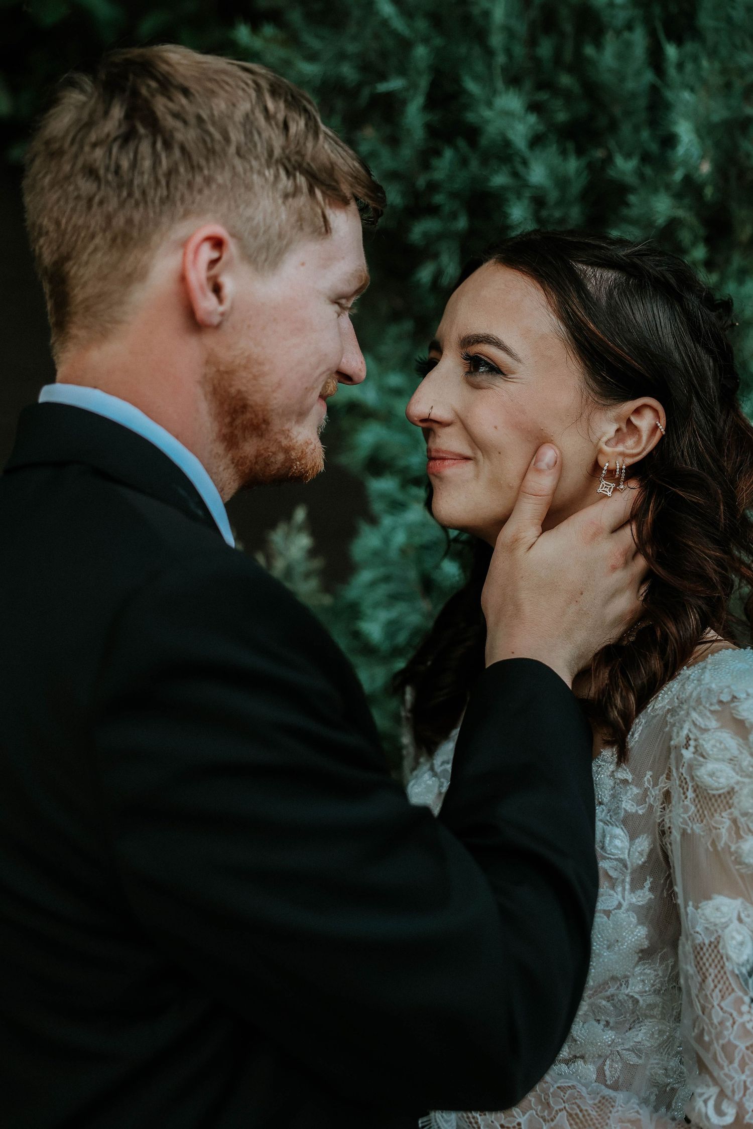 A groom tenderly cups his bride's face as they share an intimate moment, both smiling, with lush greenery in the background.