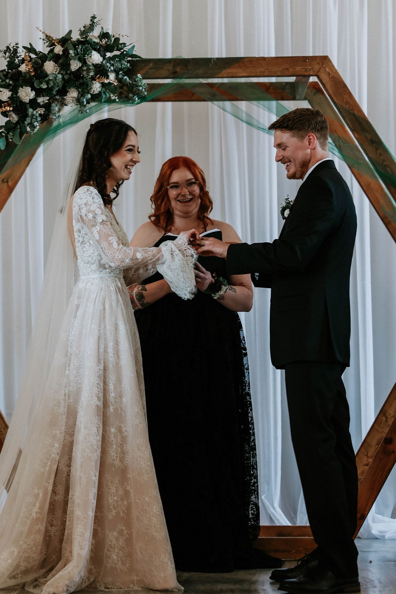 Bride and groom exchange rings at hexagonal wooden arch altar with officiant during indoor wedding ceremony.