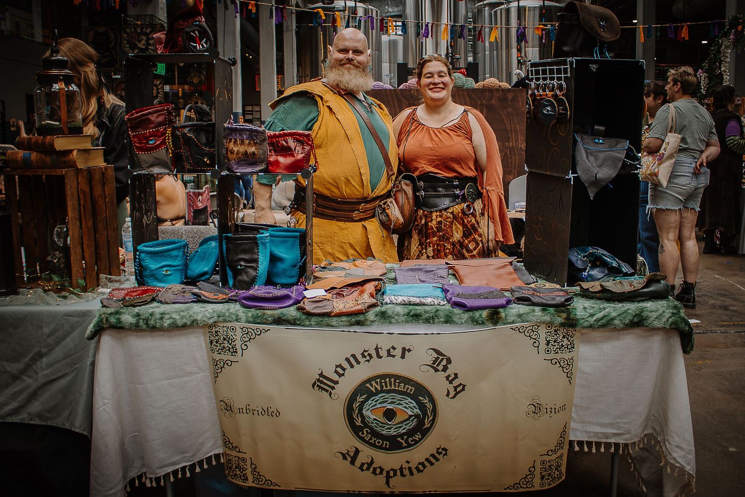 Two vendors in medieval costumes stand behind a Monster Dog Adoptions table at a fantasy market.