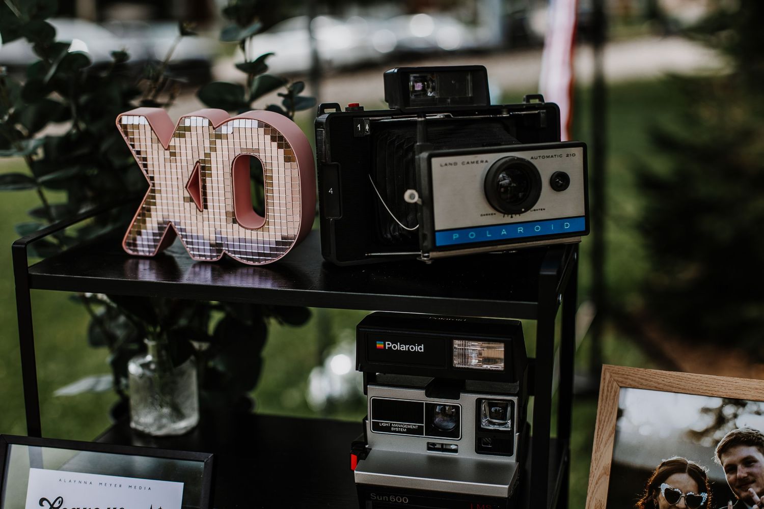Vintage Polaroid cameras and XO letter sign displayed on a black shelf at an outdoor market.