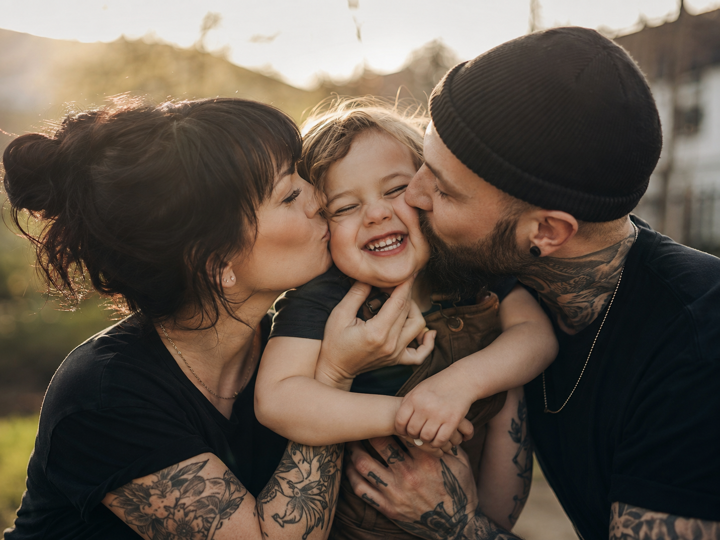 Tattooed parents kissing smiling toddler on cheeks in sunny outdoor setting.