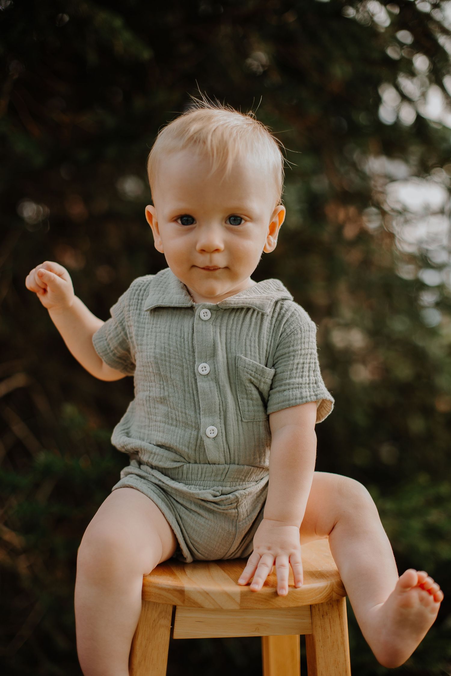 Adorable baby boy in sage green outfit sitting on wooden stool outdoors in natural light.