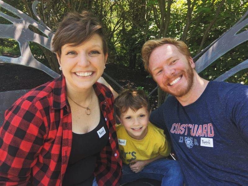Family of three smiling together for a selfie outdoors surrounded by lush green trees.