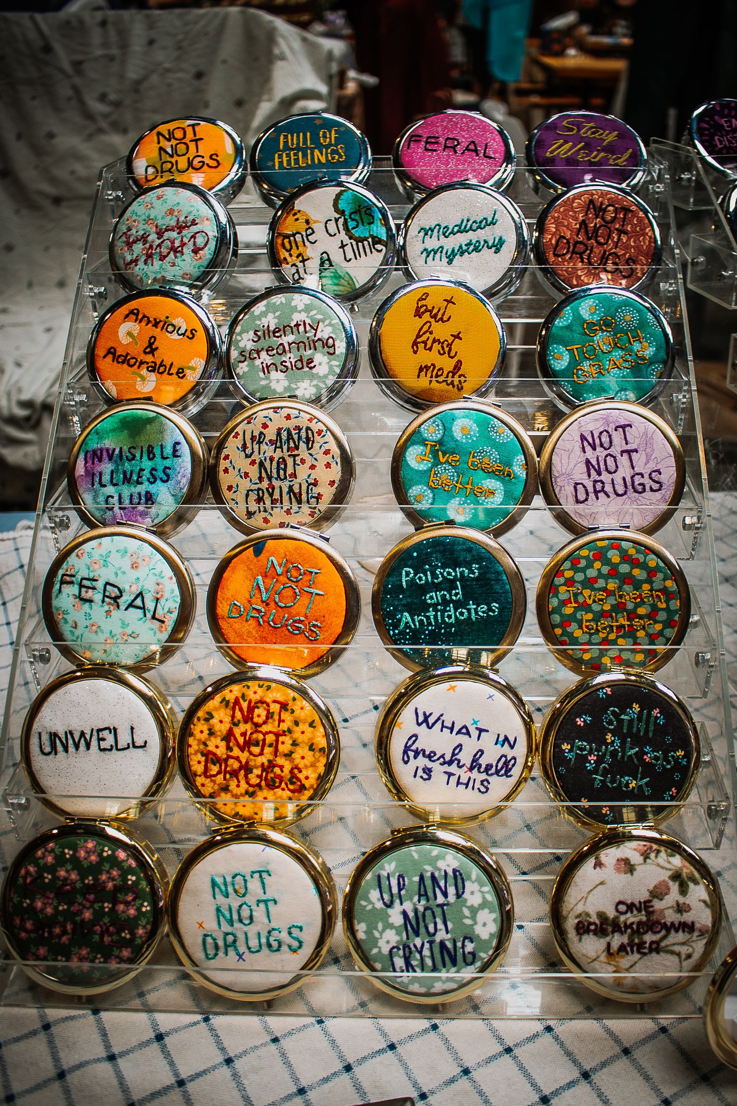 Colorful handmade pin buttons with humorous mental health phrases displayed on a clear acrylic rack at a craft market.