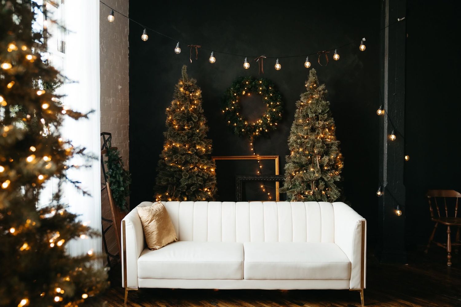 White sofa in a festive room with decorated Christmas trees, string lights, and a wreath on a dark wall.