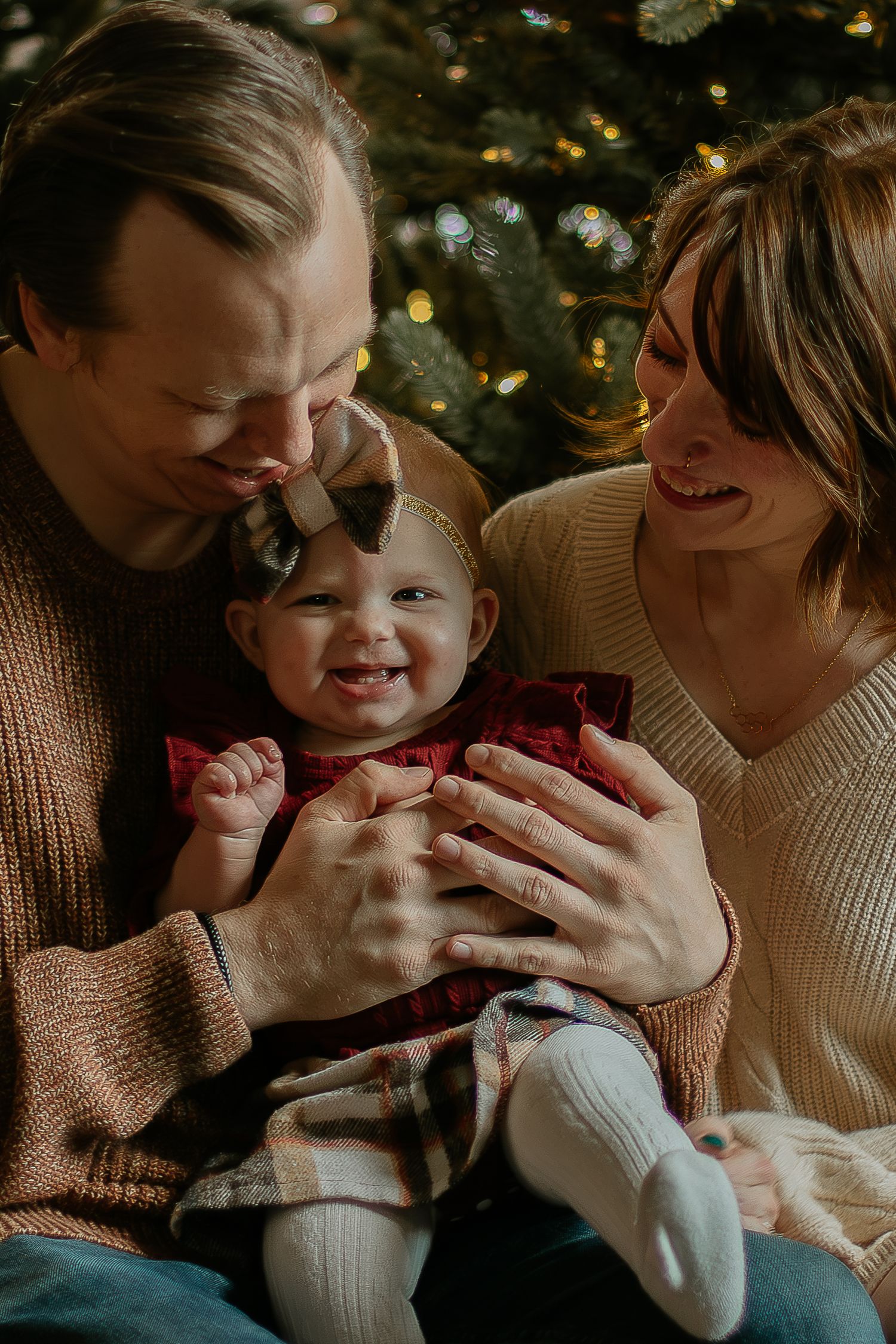Happy parents smile at laughing baby girl in red dress before Christmas tree lights.
