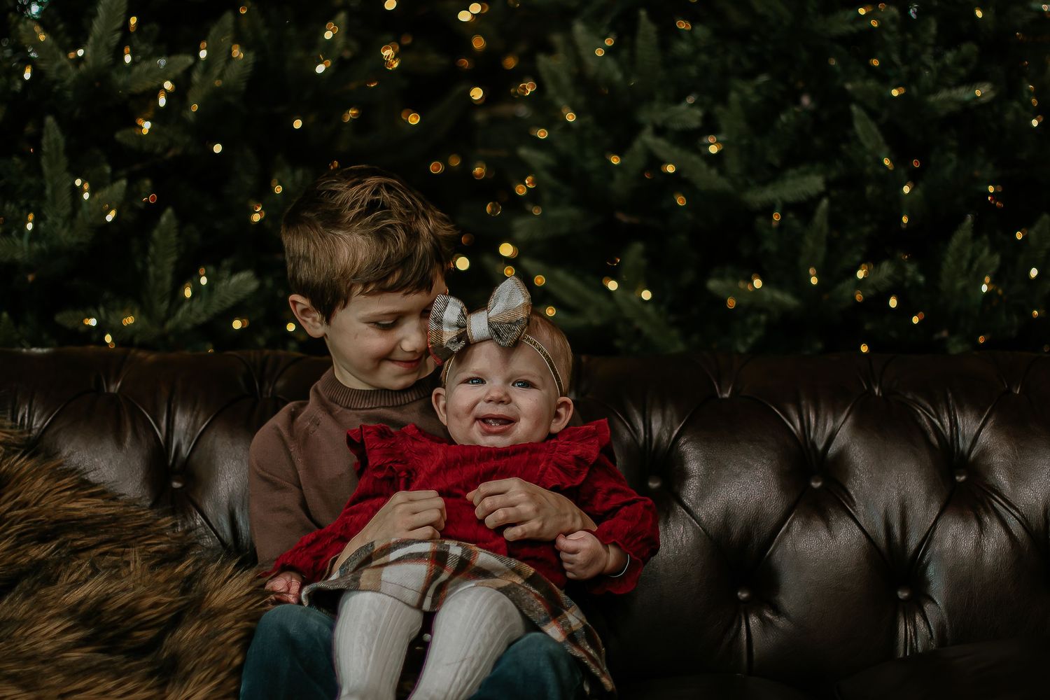 Sweet siblings pose together on leather sofa in front of glowing Christmas tree bokeh.