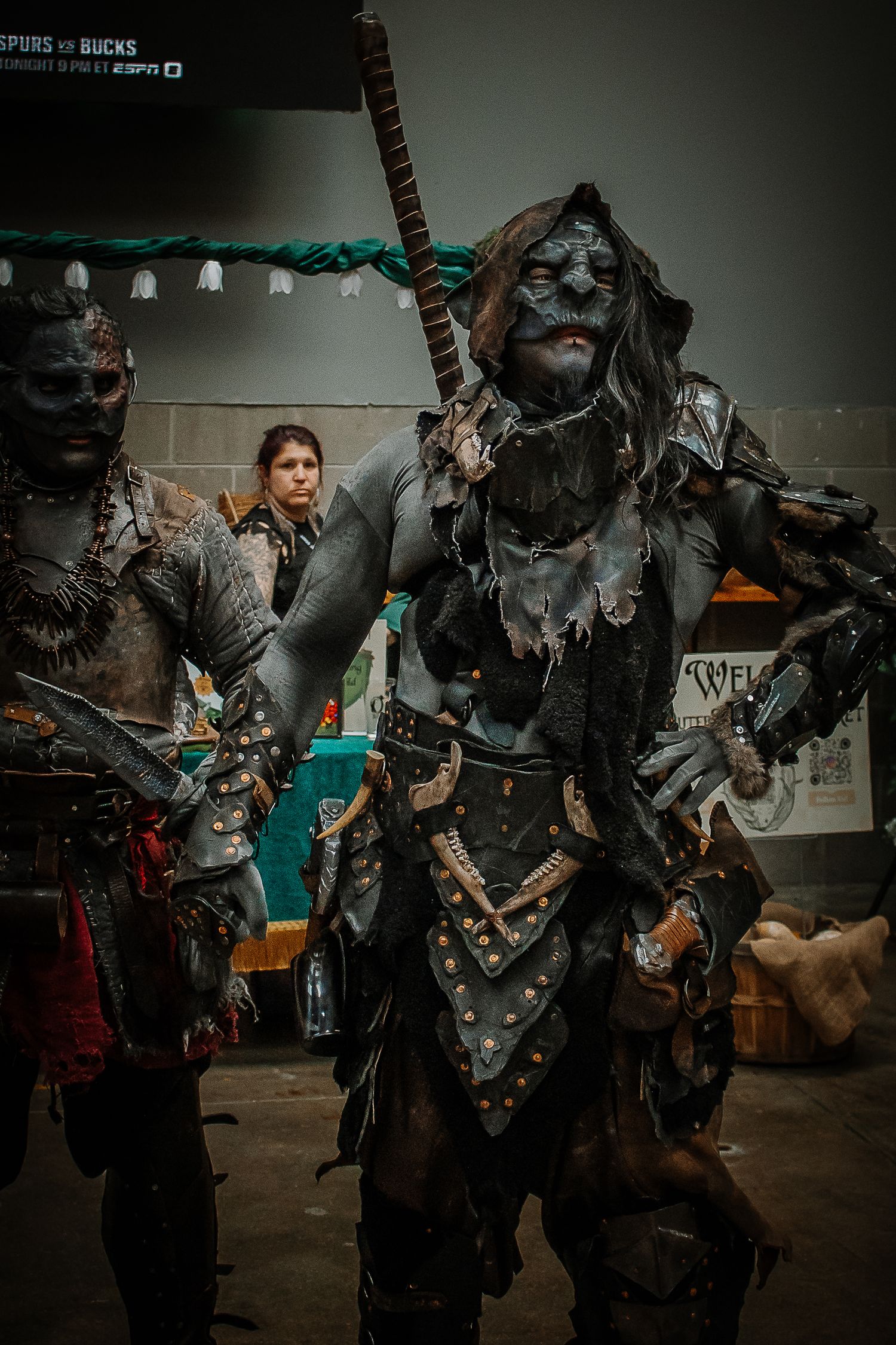 Detailed orc cosplayer in dark armor and blue body paint carries a weapon at a fantasy convention.