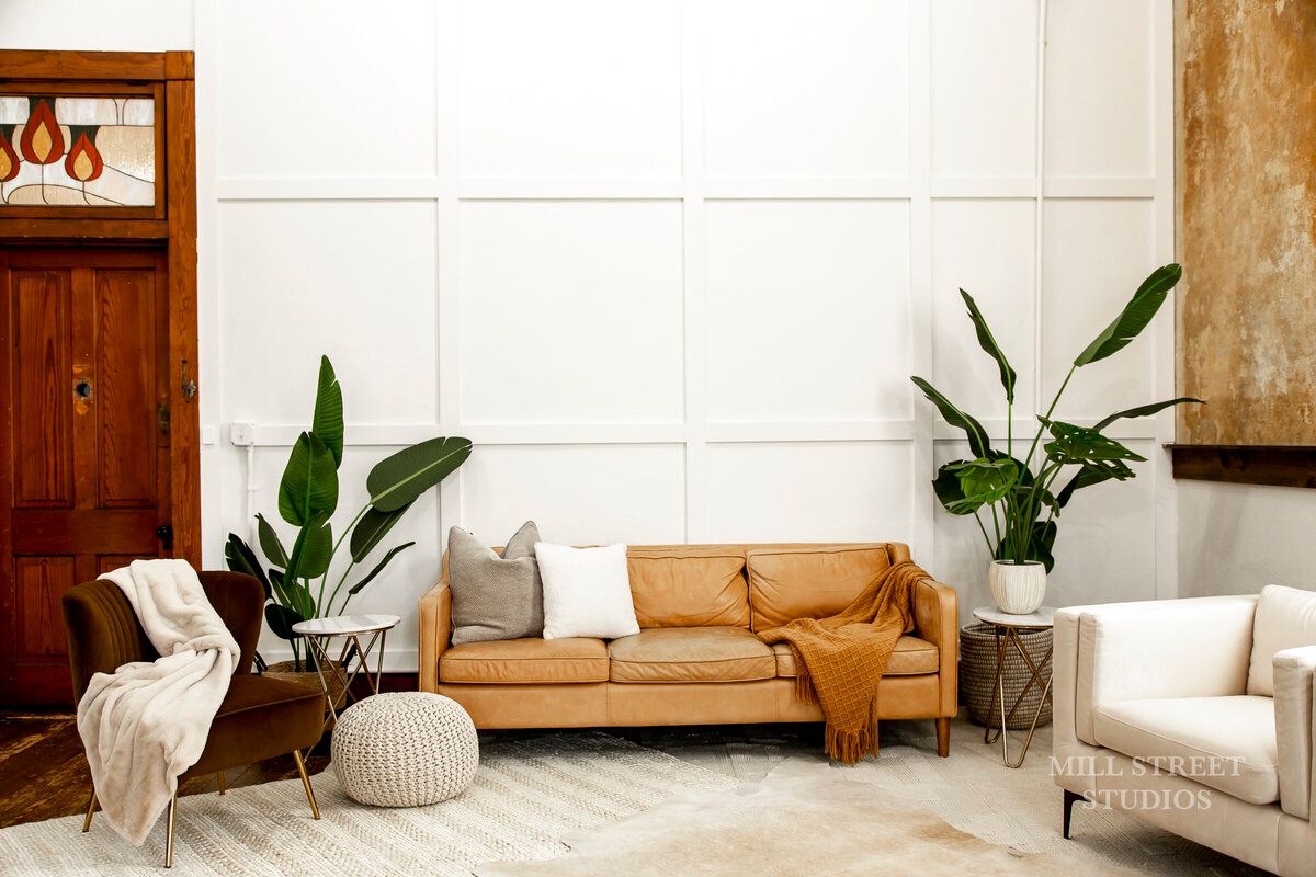Warm boho living area with caramel leather sofa, white armchair, tropical plants, and wainscoting.