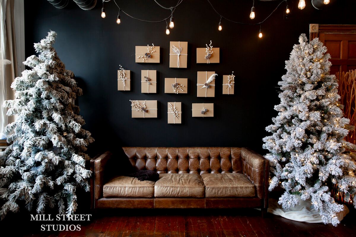Festive studio with two flocked Christmas trees flanking a brown leather tufted sofa against a dark wall with framed art.