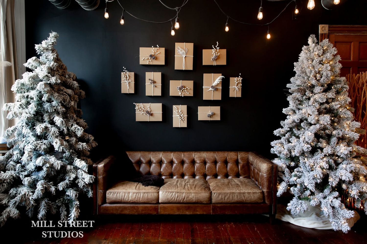 Festive studio with two flocked Christmas trees flanking a brown leather tufted sofa against a dark wall.