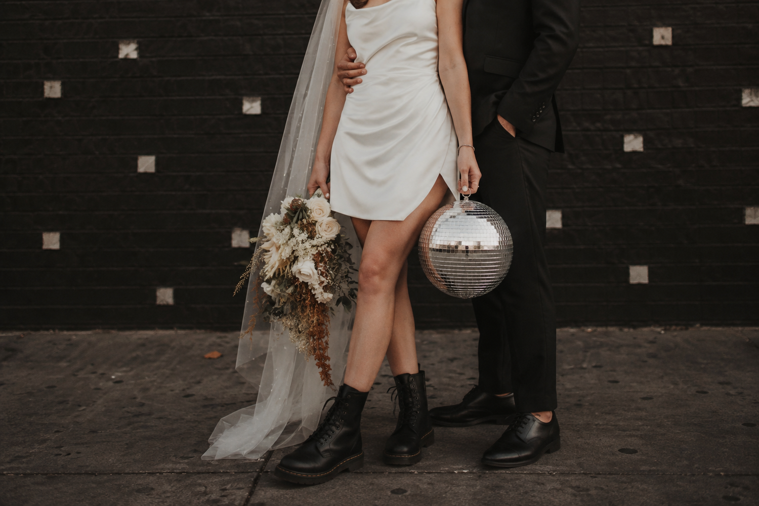 Bride in white mini dress with veil and combat boots holds bouquet beside groom holding disco ball against black brick wall.