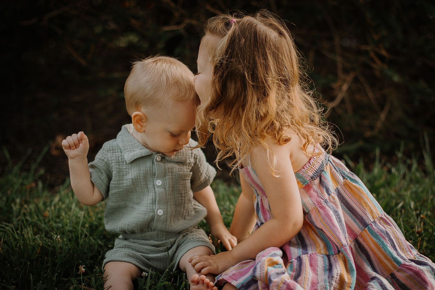 Two young children, a baby boy and a curly-haired girl in a colorful dress, sit together on green grass outdoors.