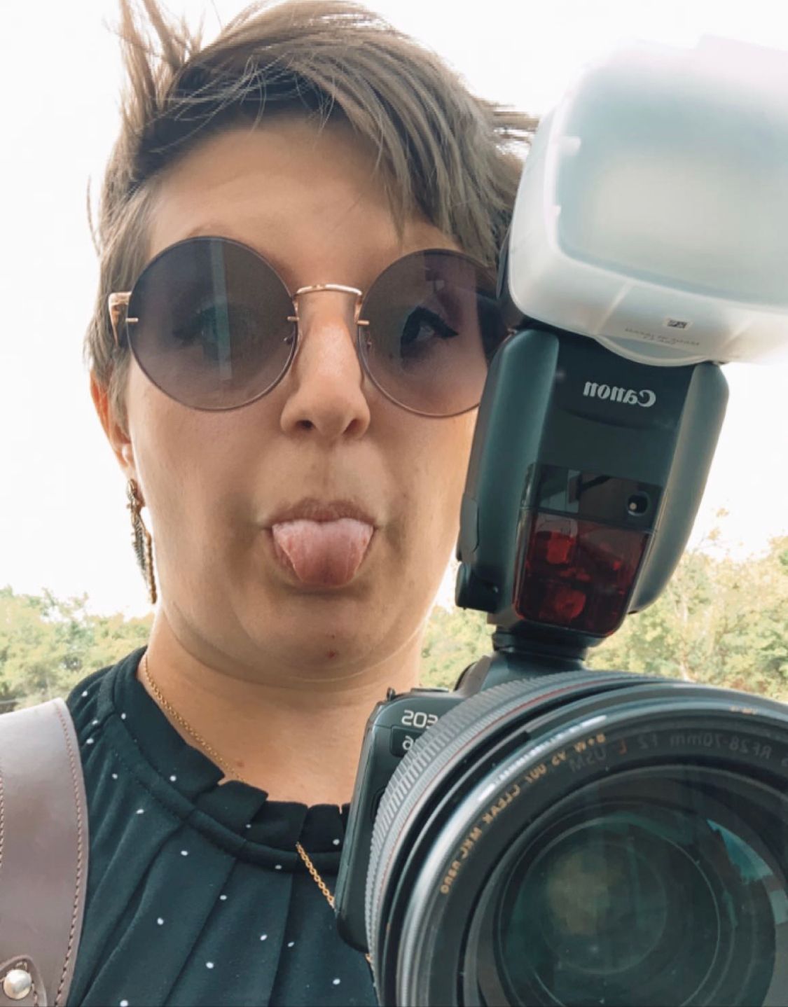 Woman sticking tongue out while holding Canon camera with flash outdoors.