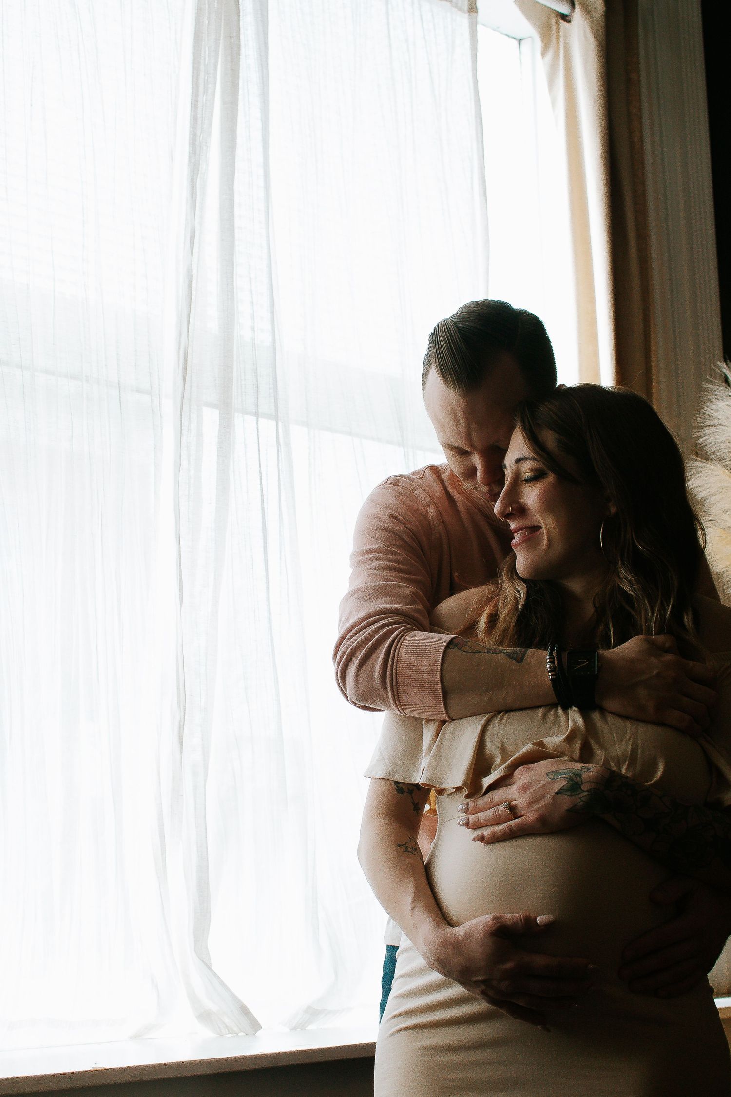 A man tenderly embraces a pregnant woman from behind near a bright window, both smiling softly.