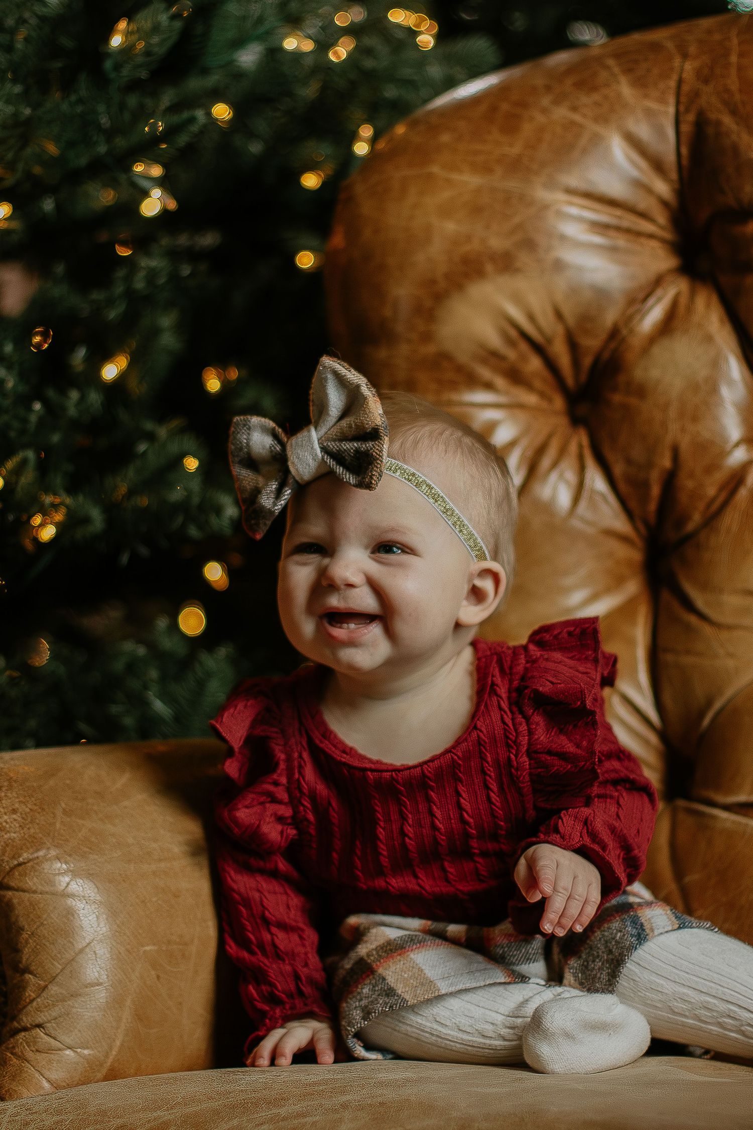 Smiling baby girl in red knit outfit sits in leather chair by Christmas tree.
