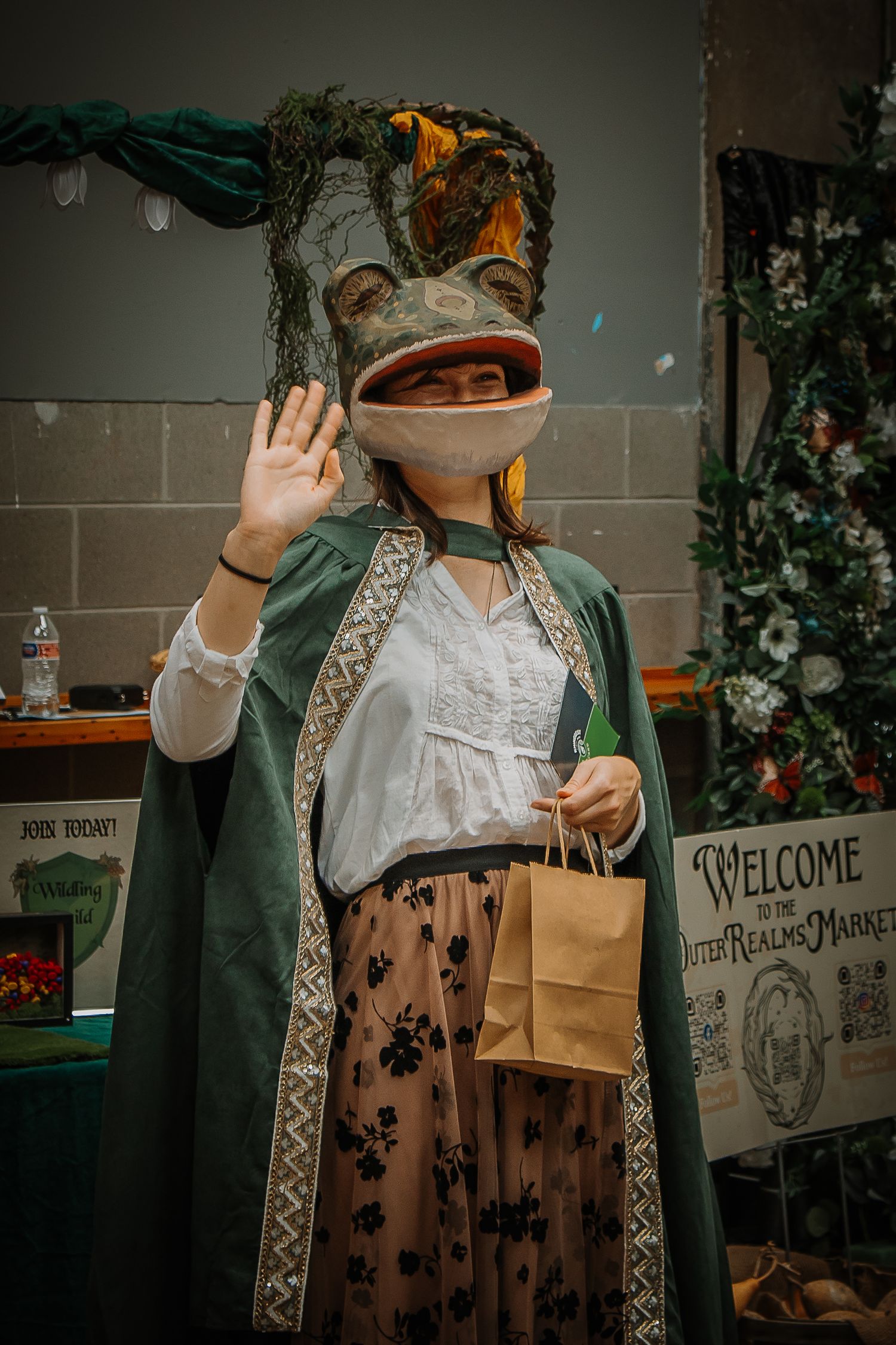 Person in green cape and frog head mask waves while holding a bag at the Otter Realms Market.