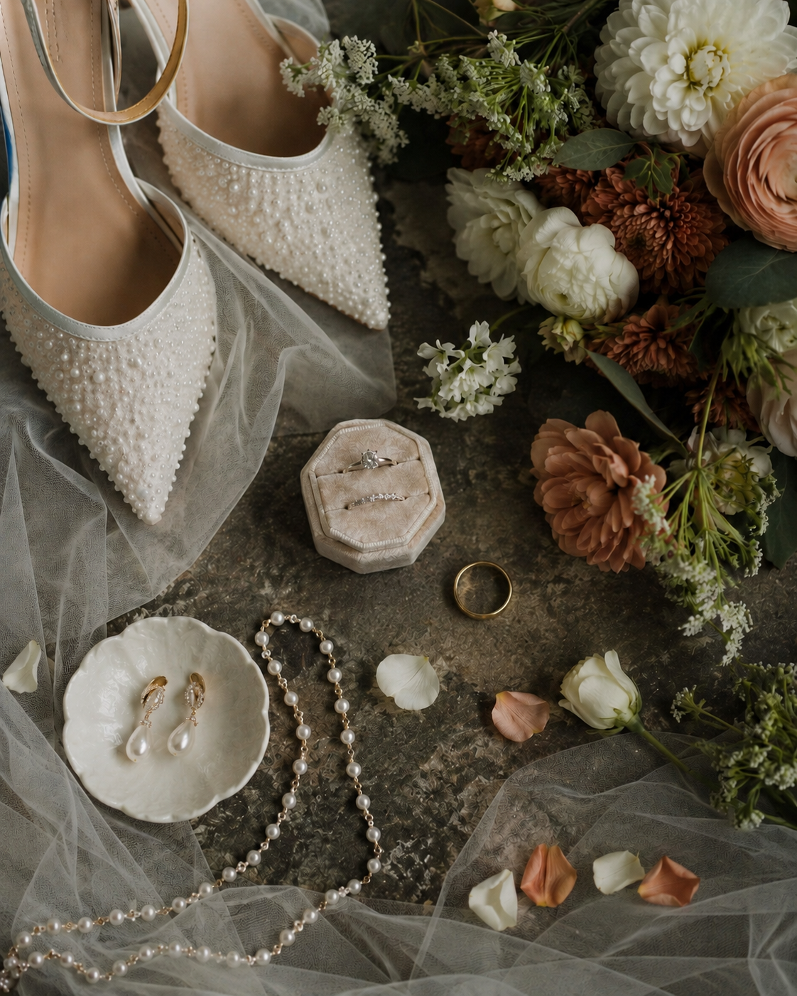 Elegant bridal flat lay with pearl heels, floral bouquet, wedding rings, pearl necklace, and earrings on a vintage surface.