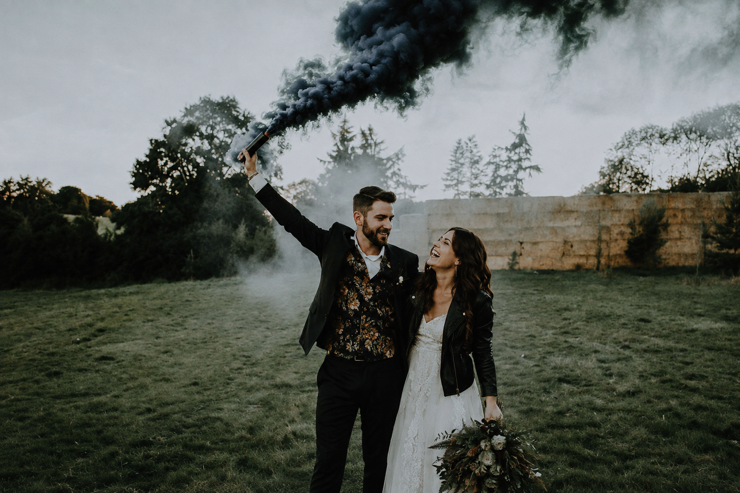 A couple celebrates their wedding outdoors, groom holding black smoke flare, bride carrying bouquet in a field.