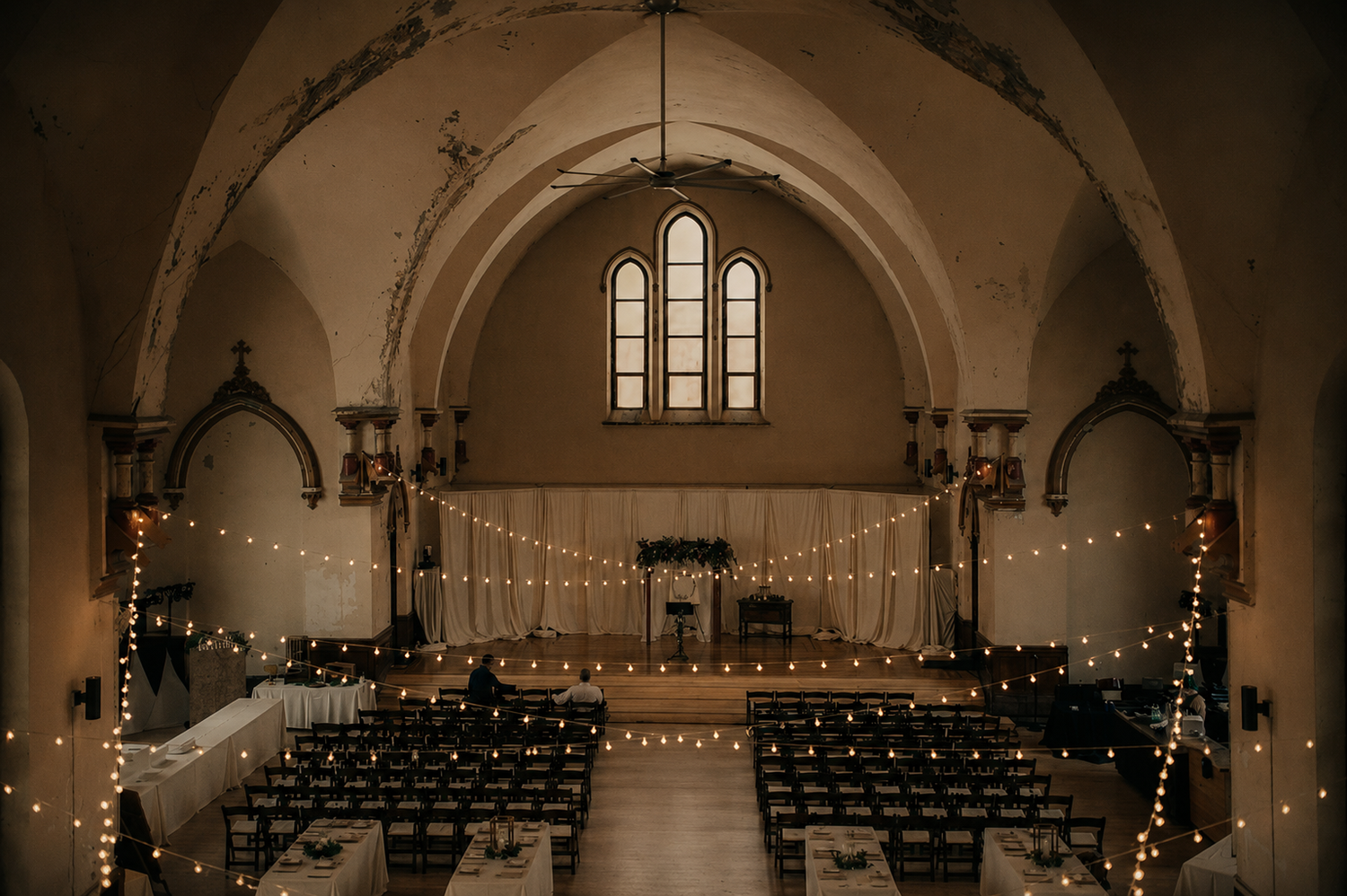 Elegant church wedding venue with string lights draped across vaulted ceilings and rows of chairs along a white aisle.
