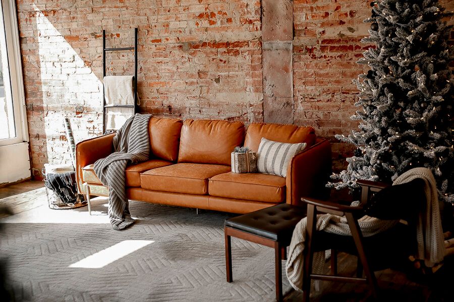 Cozy living room with a tan leather sofa, brick wall, gray throw blanket, coffee table, and a decorated Christmas tree.