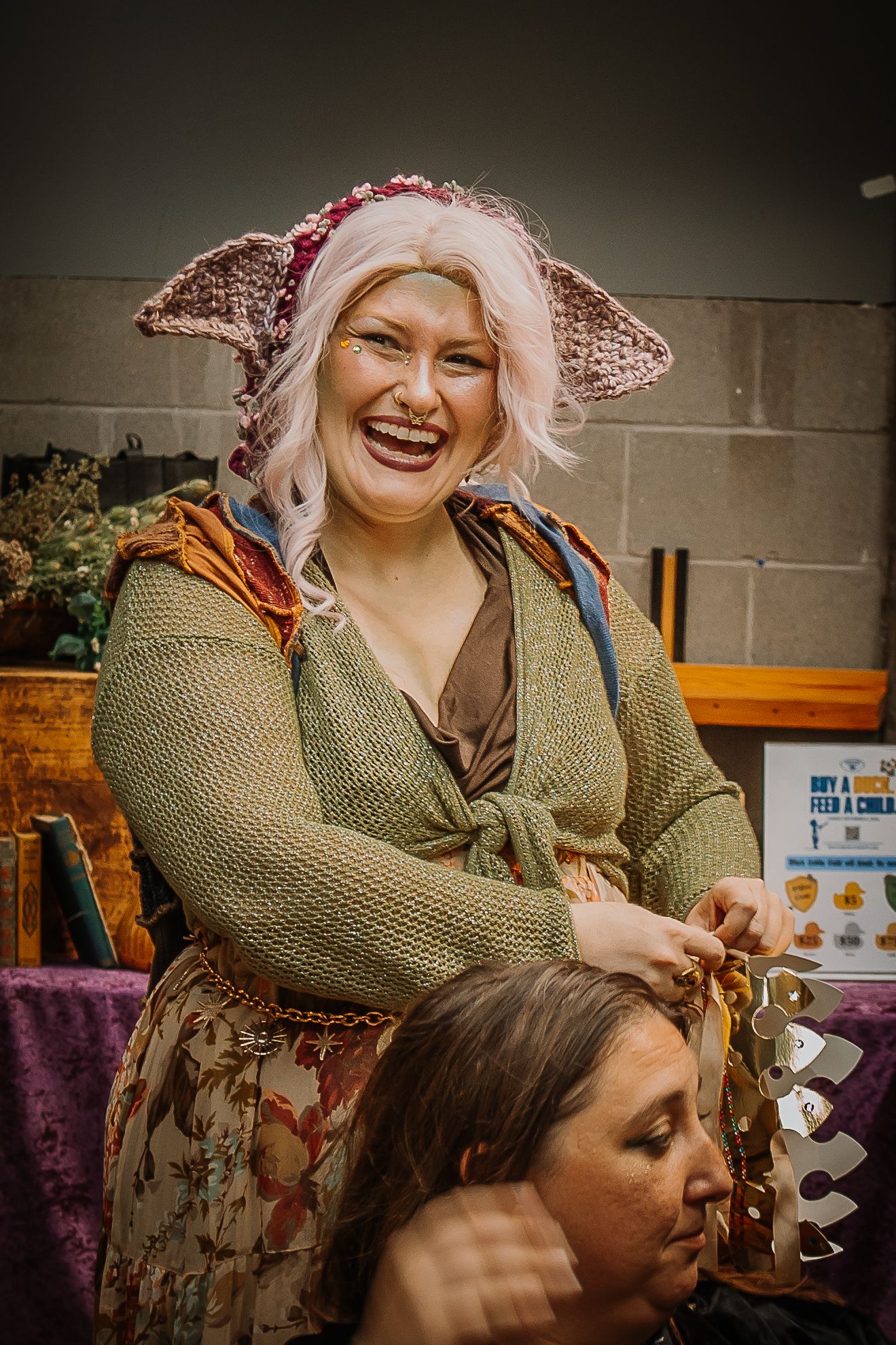Smiling woman in goblin ears and knit costume laughs while helping another person at a fantasy fair.