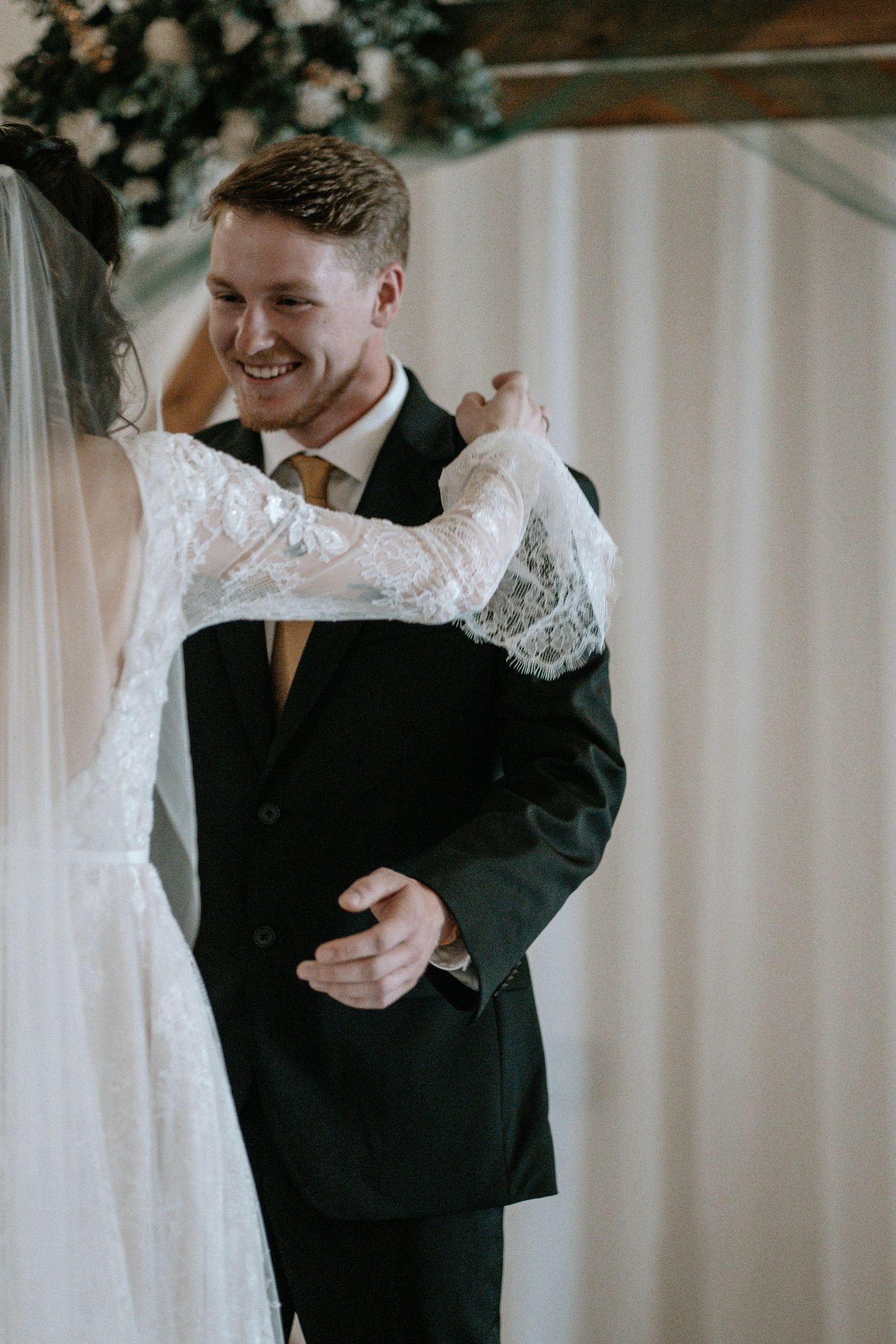 Smiling groom in black suit with gold tie embraces bride in lace wedding dress during indoor ceremony.