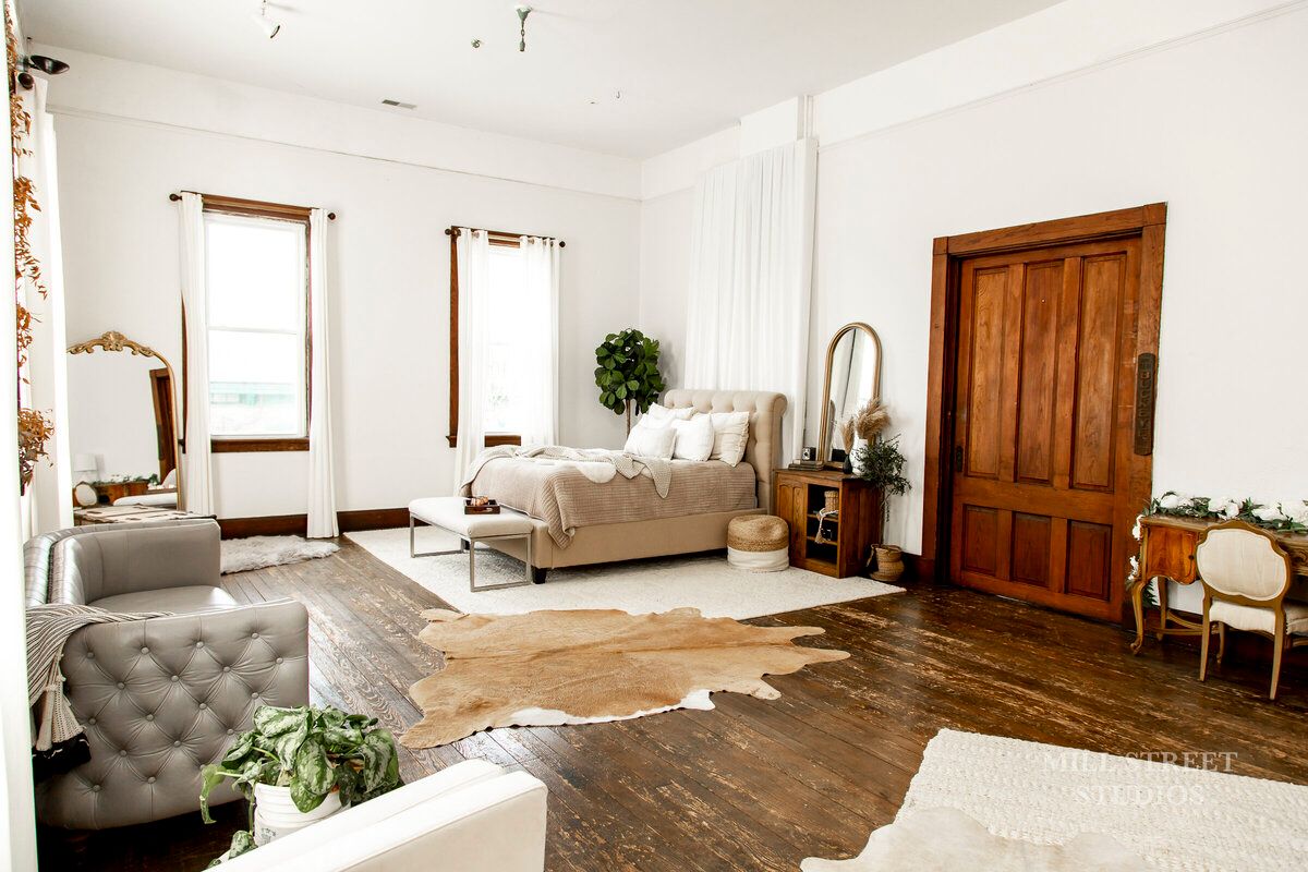Spacious boho studio with bed, gray tufted sofa, cowhide rug, wood floors, and tall wooden doors.