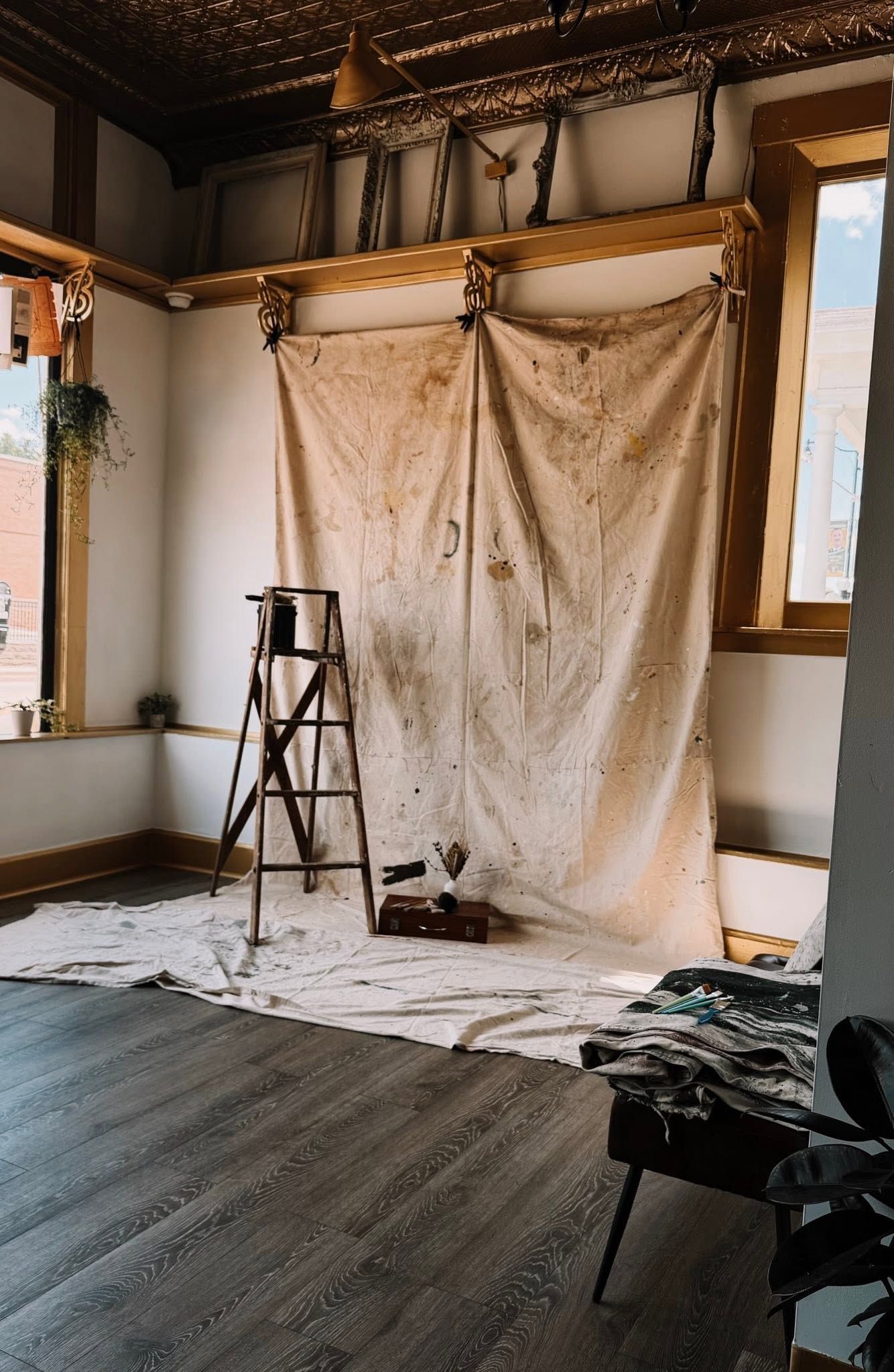 Paint-splattered drop cloth draped over wooden frame with vintage ladder in rustic studio space with ornate tin ceiling.