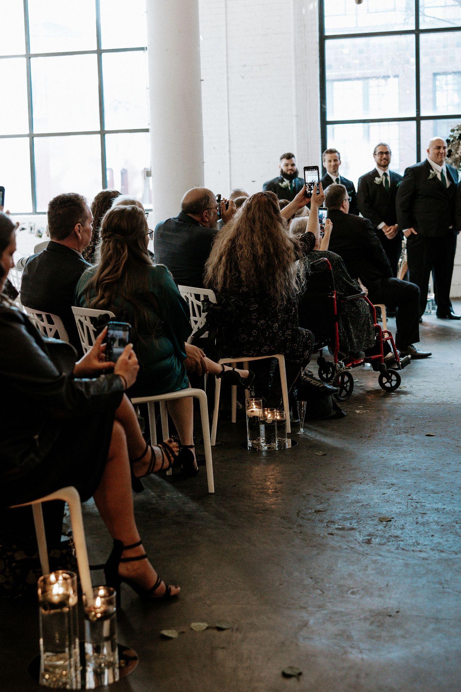 Guests seated in white chairs at an industrial wedding venue, holding phones to capture the ceremony aisle moment.