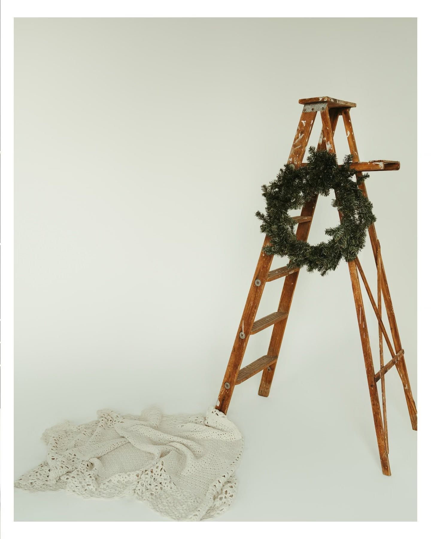 Rustic wooden ladder with a green Christmas wreath leaning against white backdrop, with lace blanket on floor.