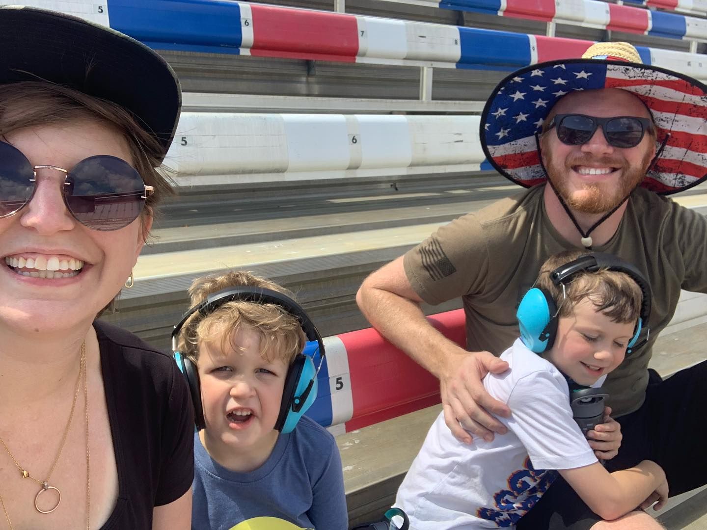 Family selfie with two young children wearing ear protection at an outdoor racing event.
