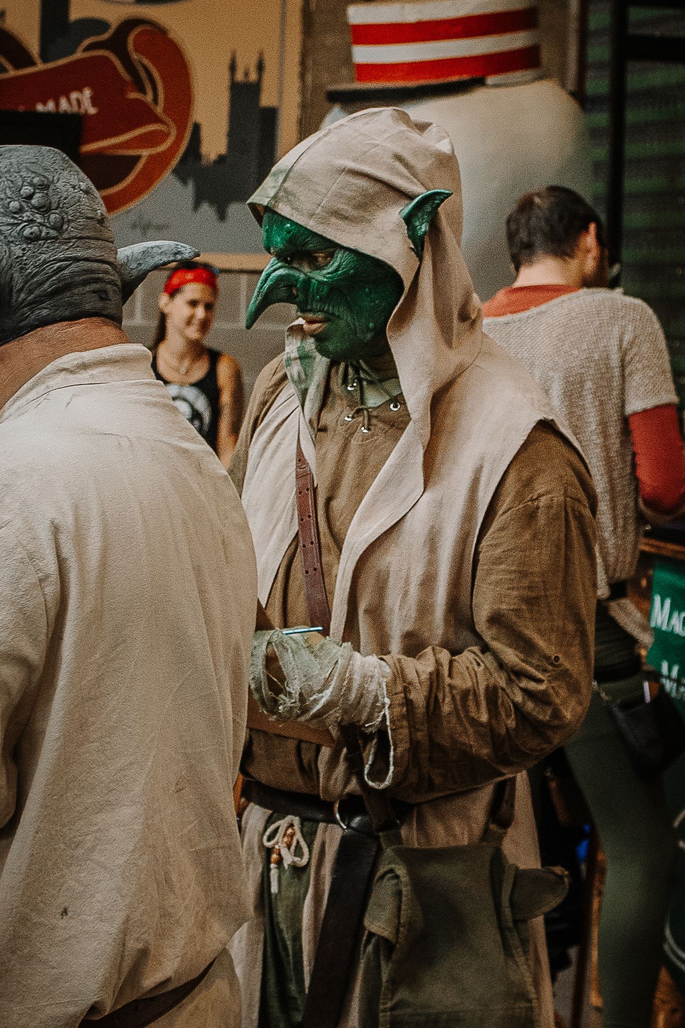 Person in elaborate green goblin costume with hooded robe at a fantasy themed market event.