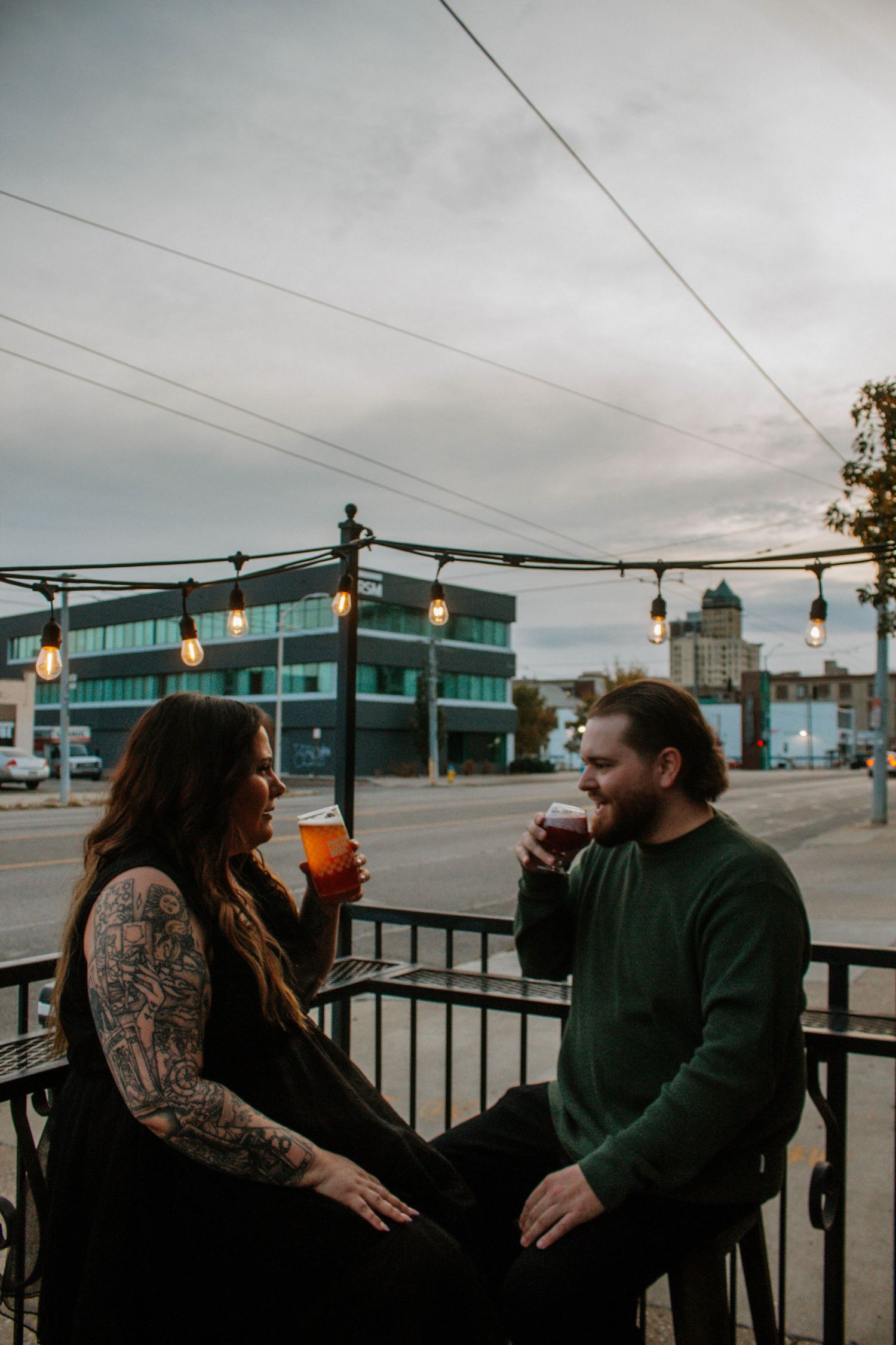Tattooed couple enjoying craft beers on an outdoor patio with city lights at dusk.