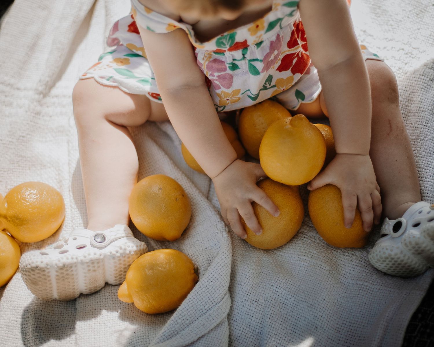Baby in floral outfit sitting on white blanket surrounded by fresh yellow lemons and oranges in natural sunlight.