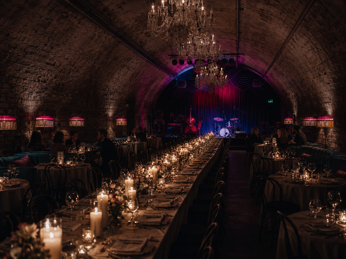 Elegant candlelit banquet hall in a stone vaulted tunnel with chandeliers and a stage glowing in purple light.