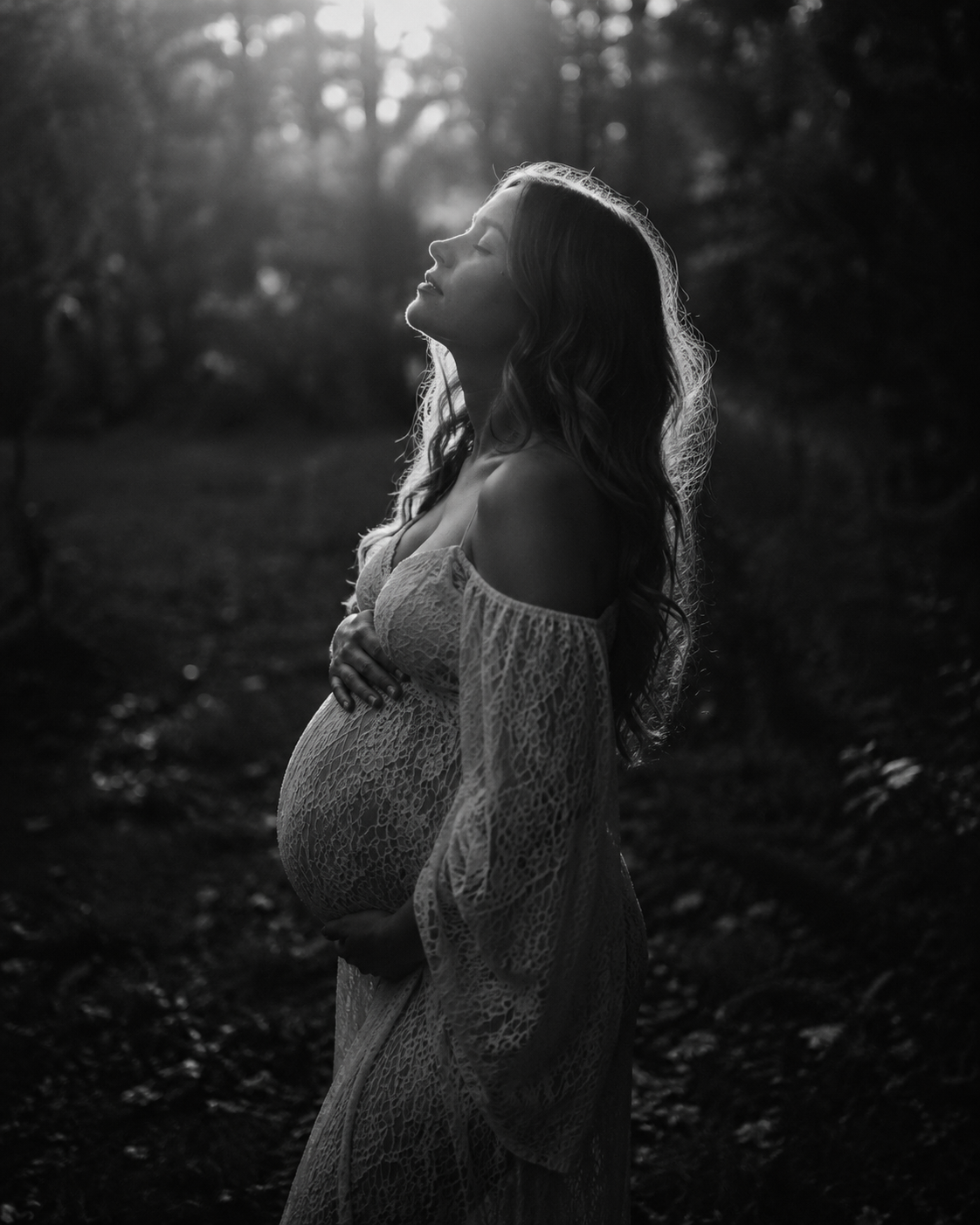 Black and white maternity photo of pregnant woman in lace dress holding belly, backlit by sunlight in forest.