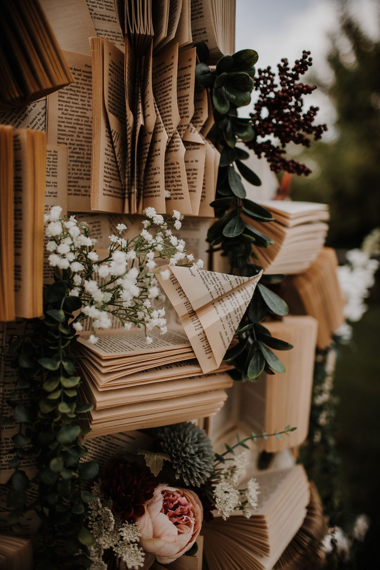 Decorative wall of folded book pages adorned with greenery, white baby's breath, and pink roses for a literary wedding.