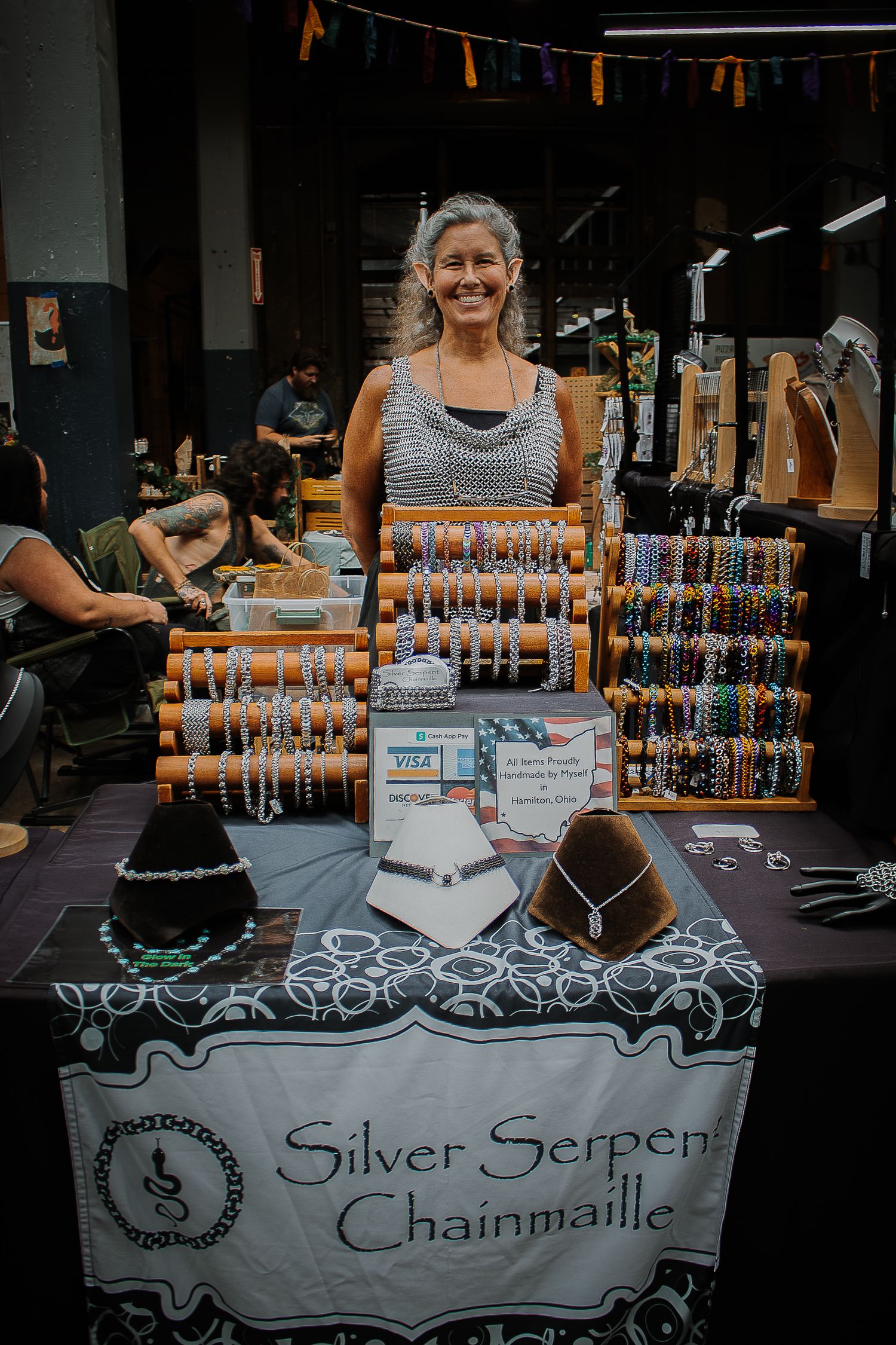 Smiling woman standing behind Silver Serpent Chainmaille jewelry booth displaying colorful beaded bracelets and necklaces.