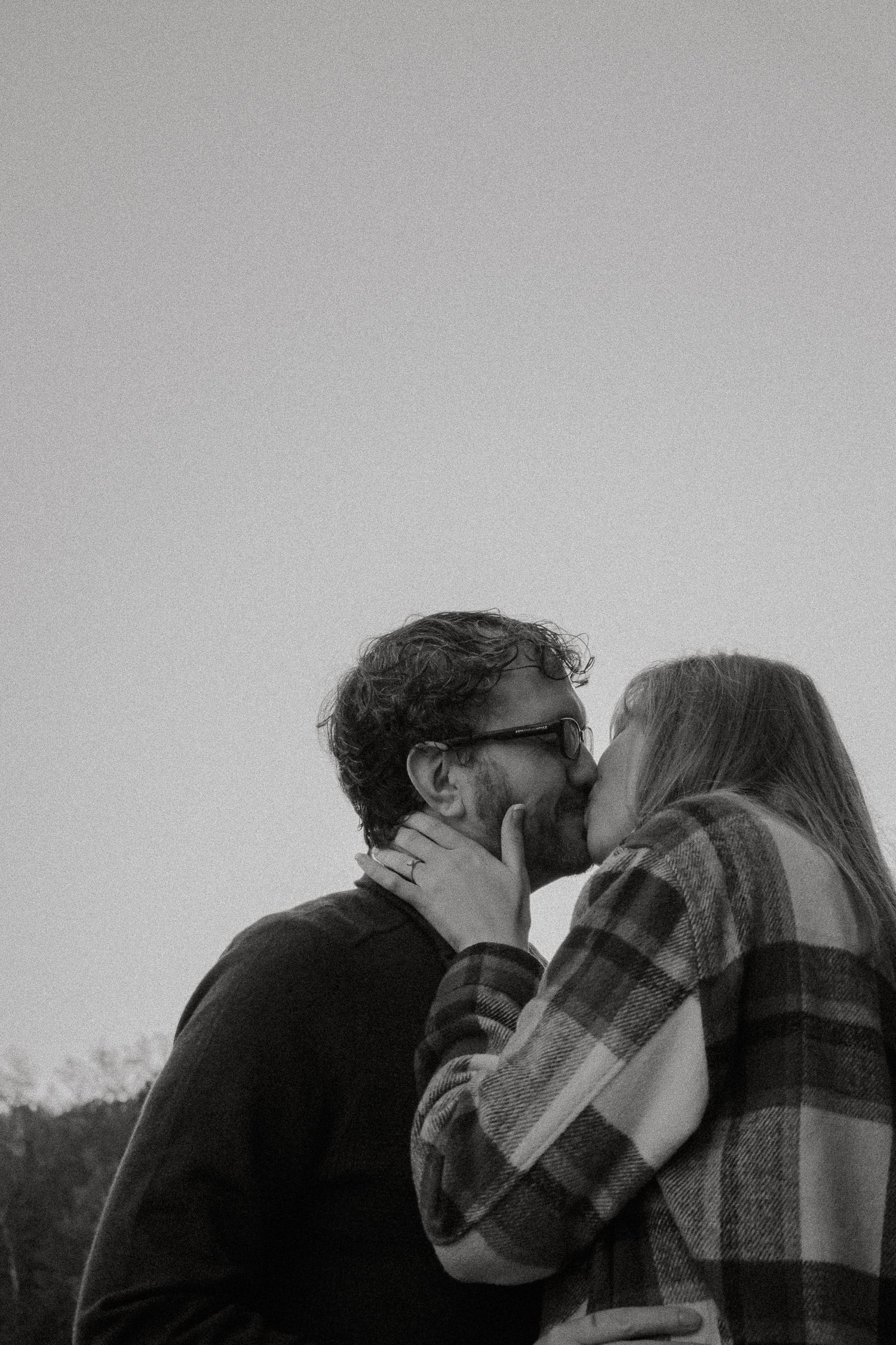 Black and white photo of a couple sharing a romantic kiss outdoors against a clear sky.