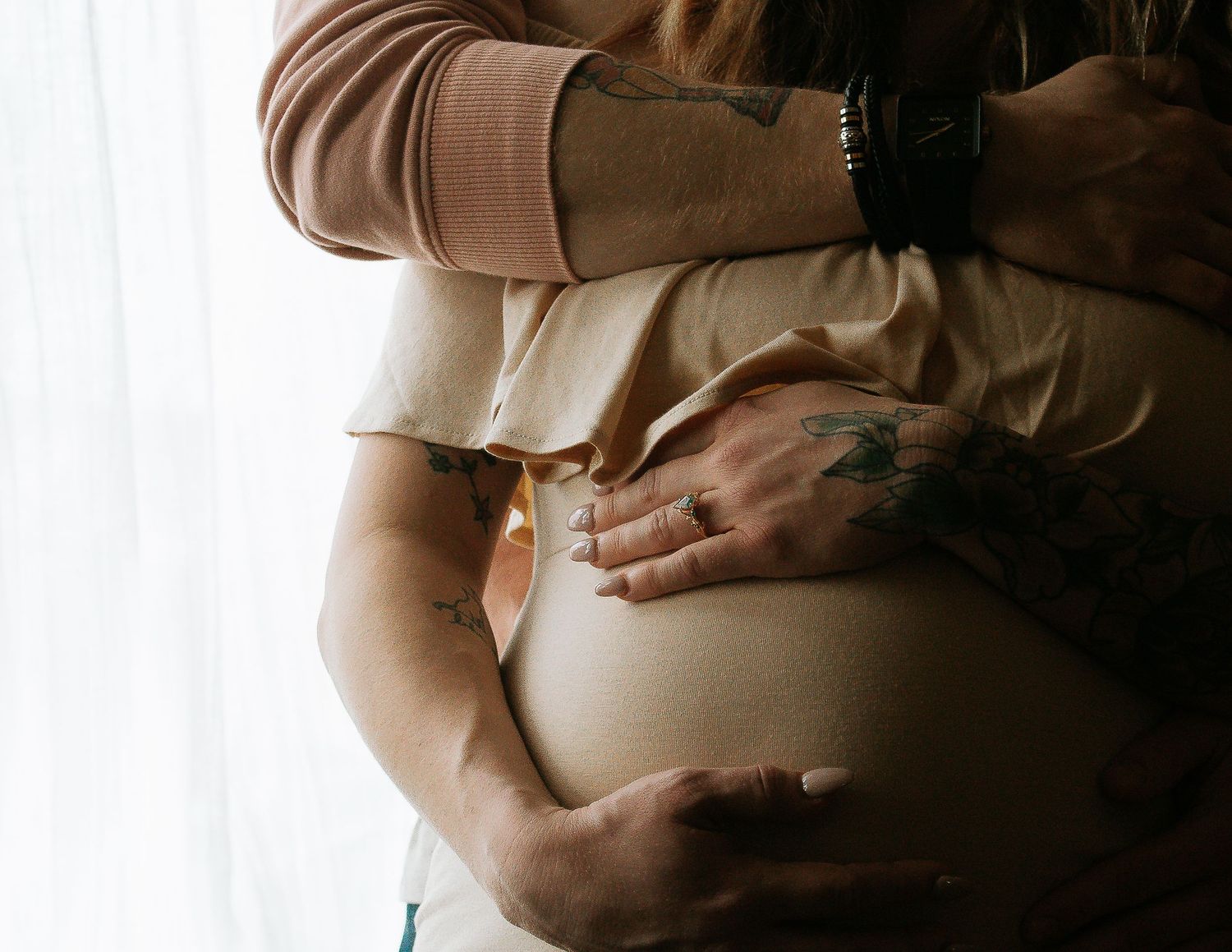 A couple with tattoos cradles a pregnant belly near a sheer curtained window in soft light.