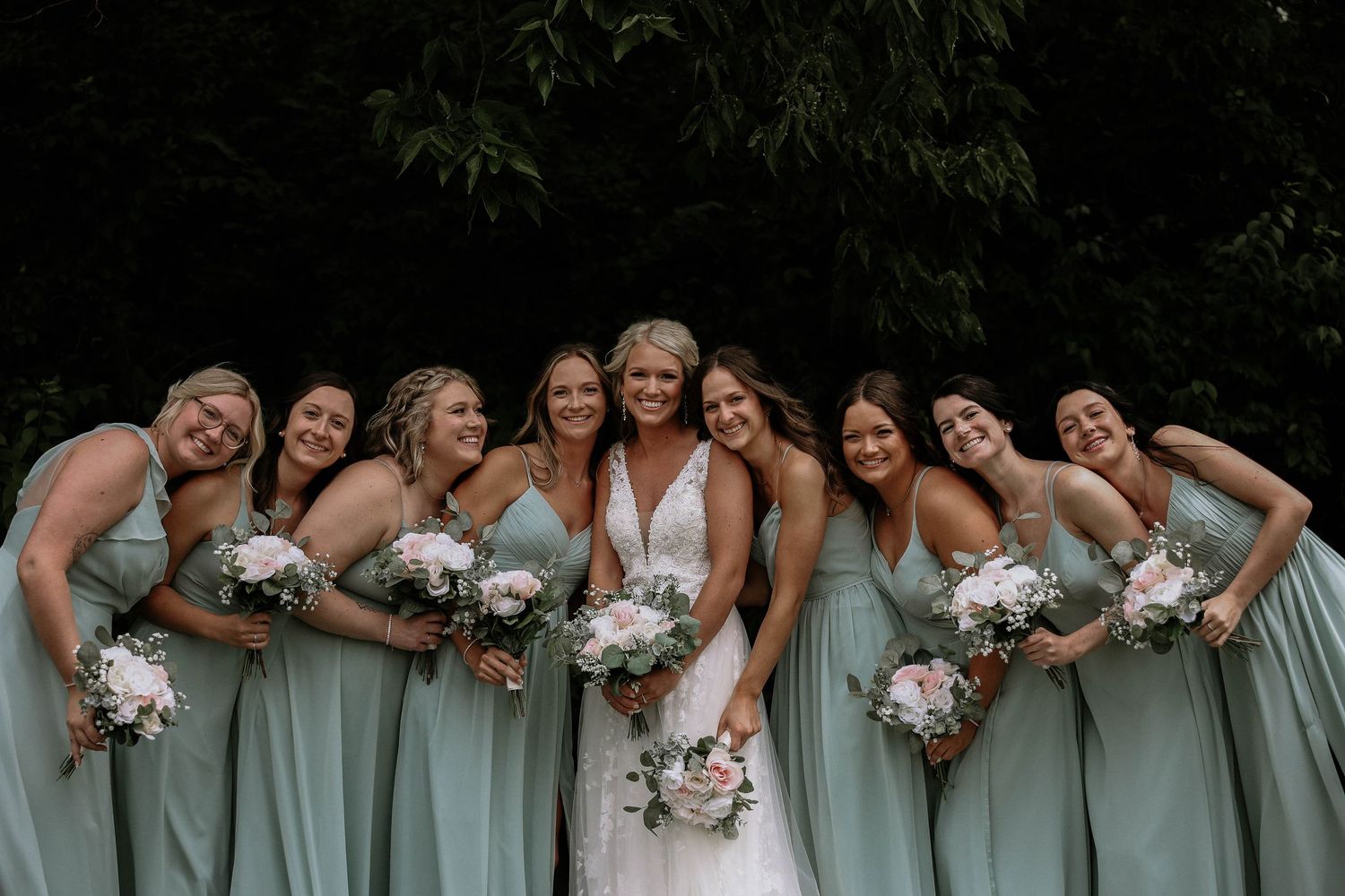 Bride in white gown poses with eight bridesmaids in sage green dresses holding white floral bouquets outdoors.